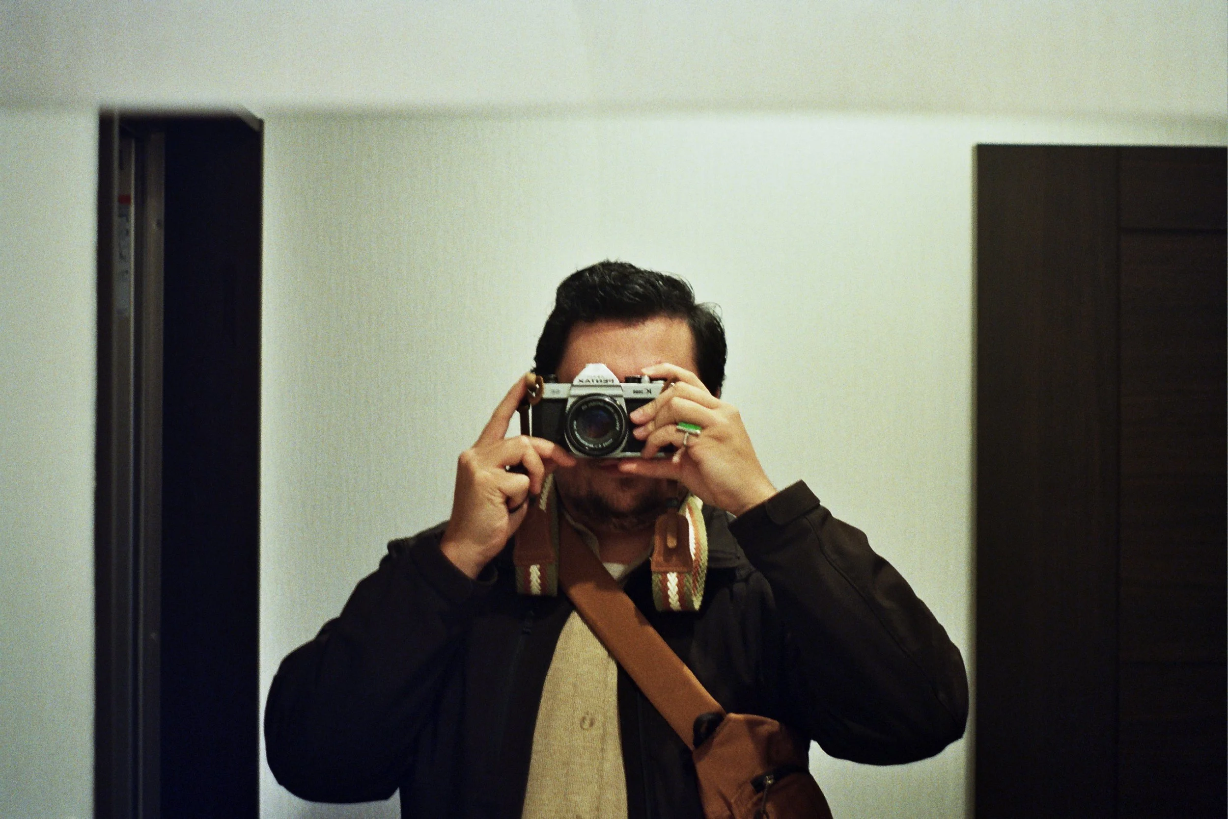 Person taking a selfie in front of a mirror with a camera, wearing a black jacket, beige sweater, and green ring, carrying a brown bag strap.