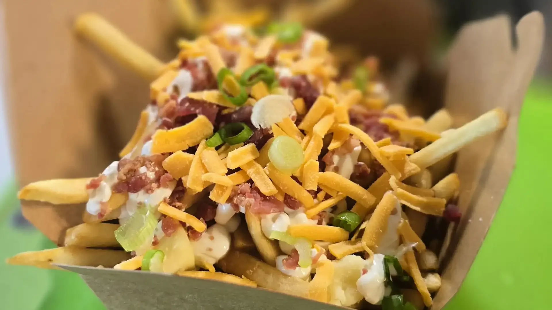 Loaded French fries topped with shredded cheese, chopped green onions, bacon bits, and sour cream in a paper food tray.