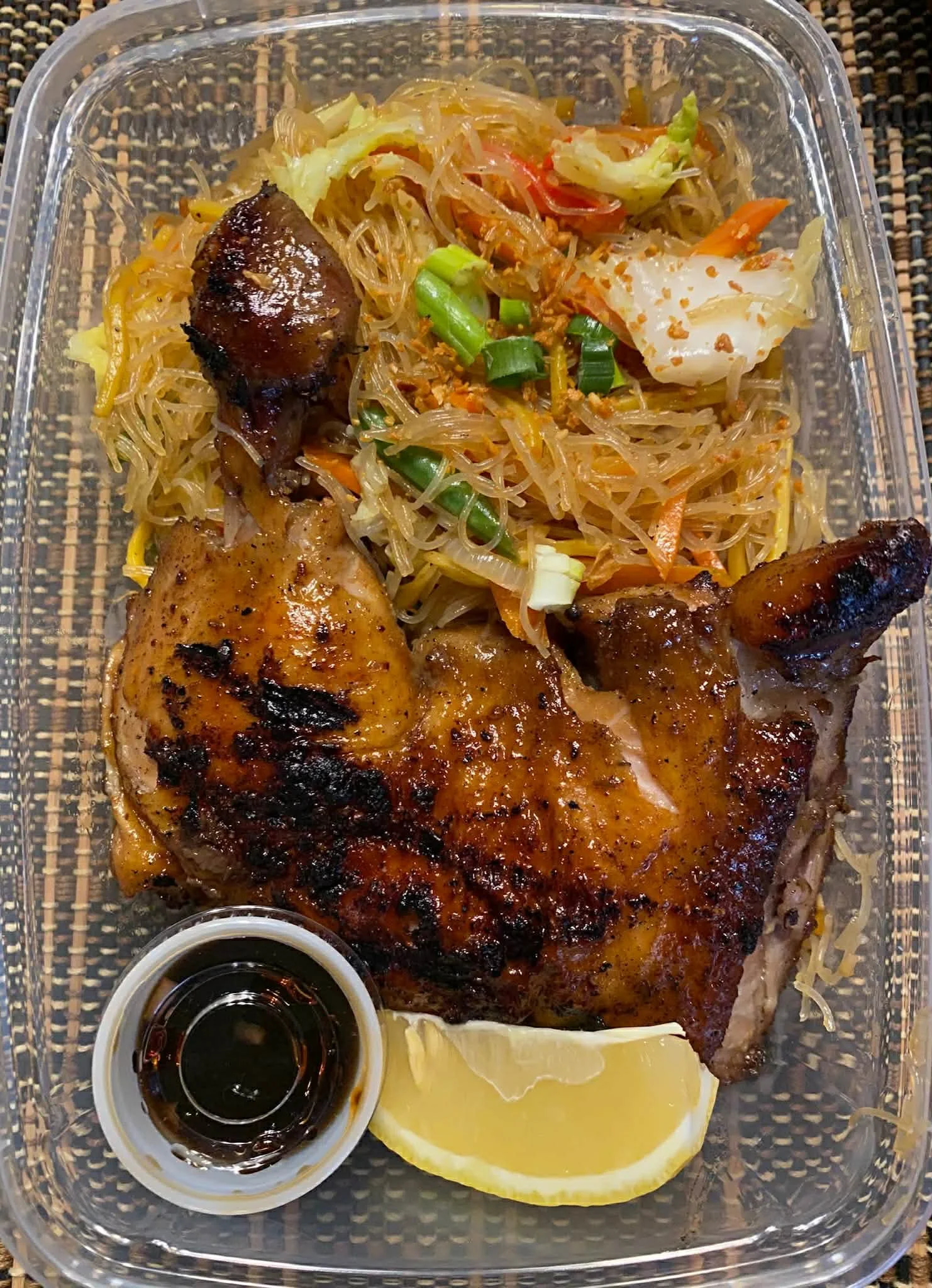 Grilled chicken leg quarter with a lemon wedge, a small cup of soy sauce, and a side of stir-fried glass noodles with vegetables, served in a clear plastic container on a woven placemat.