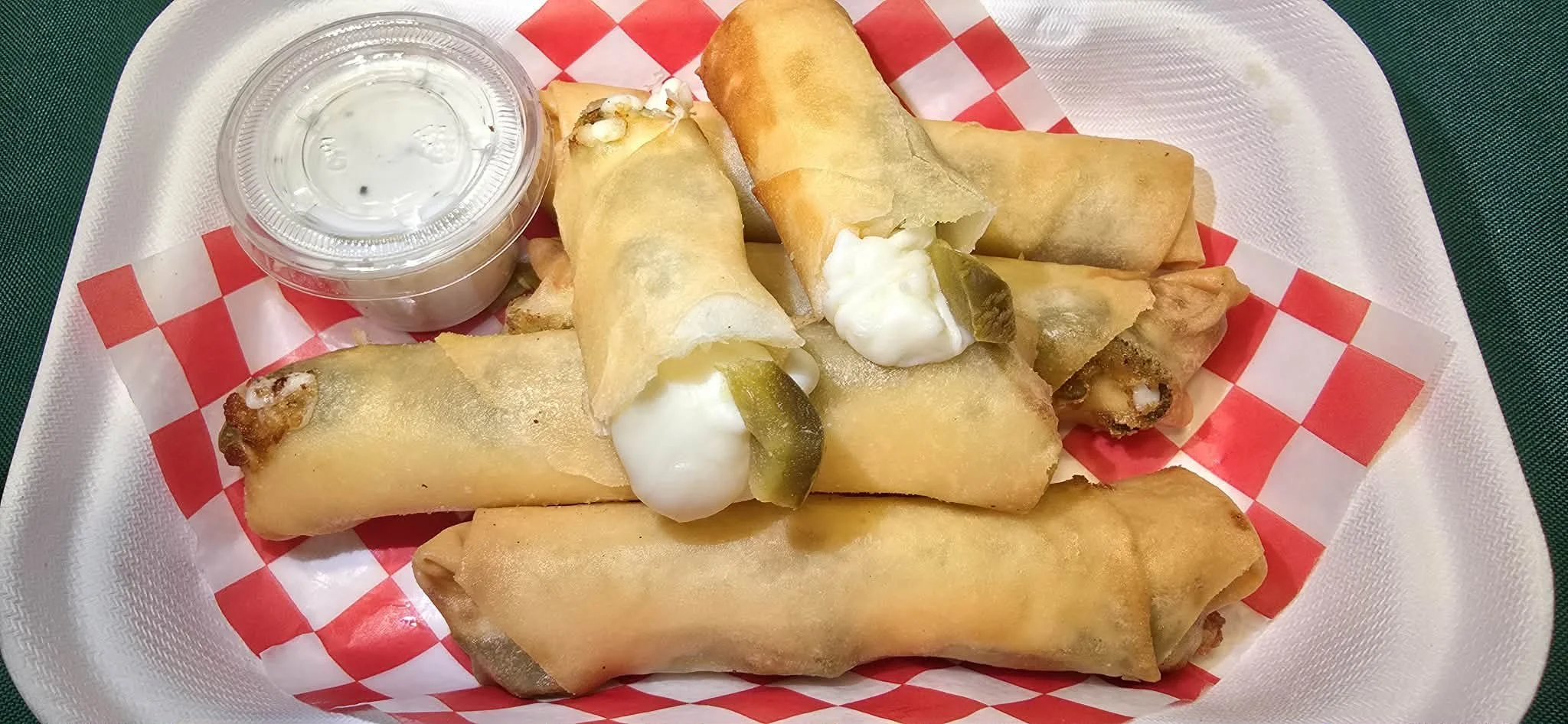Plate of five beef and cheese chimichangas with sour cream, pickles, and a small cup of sauce on a red and white checkered paper in a white tray.