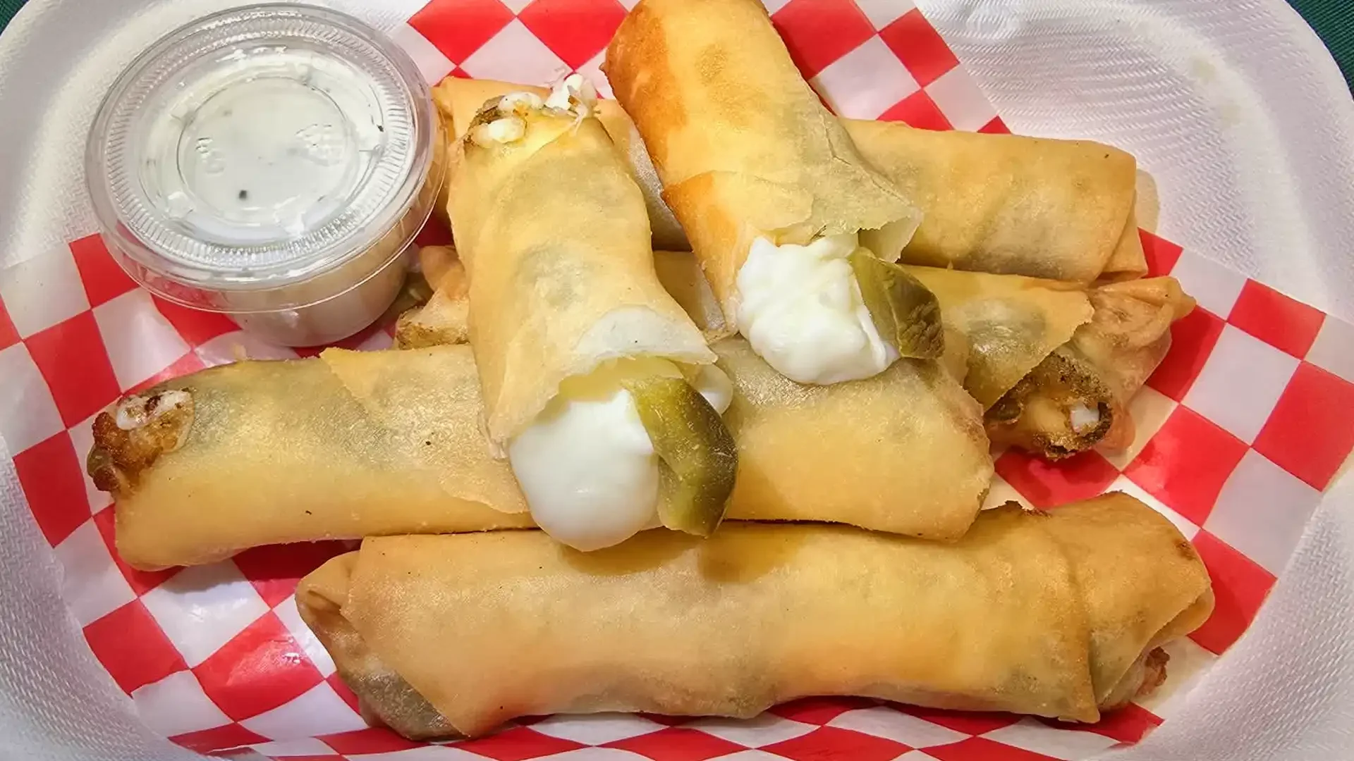 Several cheese and pickle egg rolls with dipping sauce on red and white checkered paper in a basket.