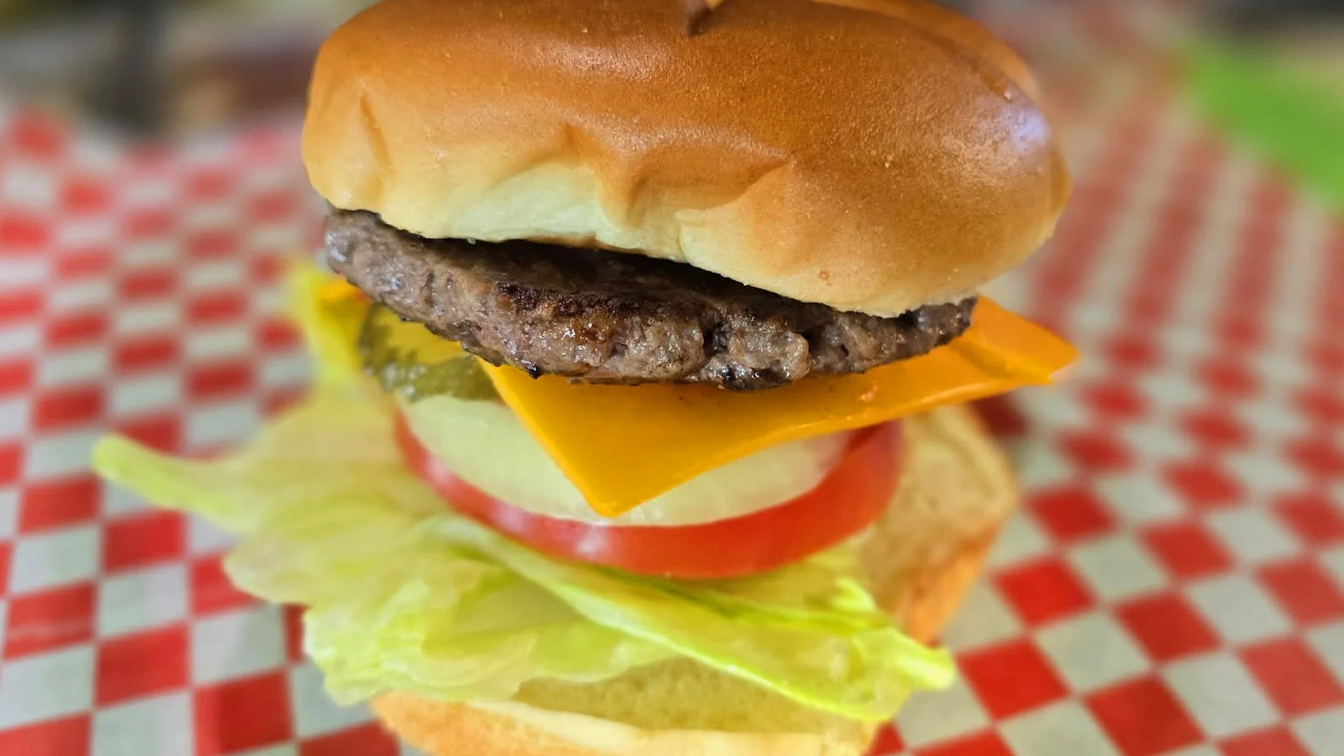 A close-up of a cheeseburger with a beef patty, cheddar cheese, lettuce, tomato, pickles, and a sandwich bun on a red and white checkered paper.