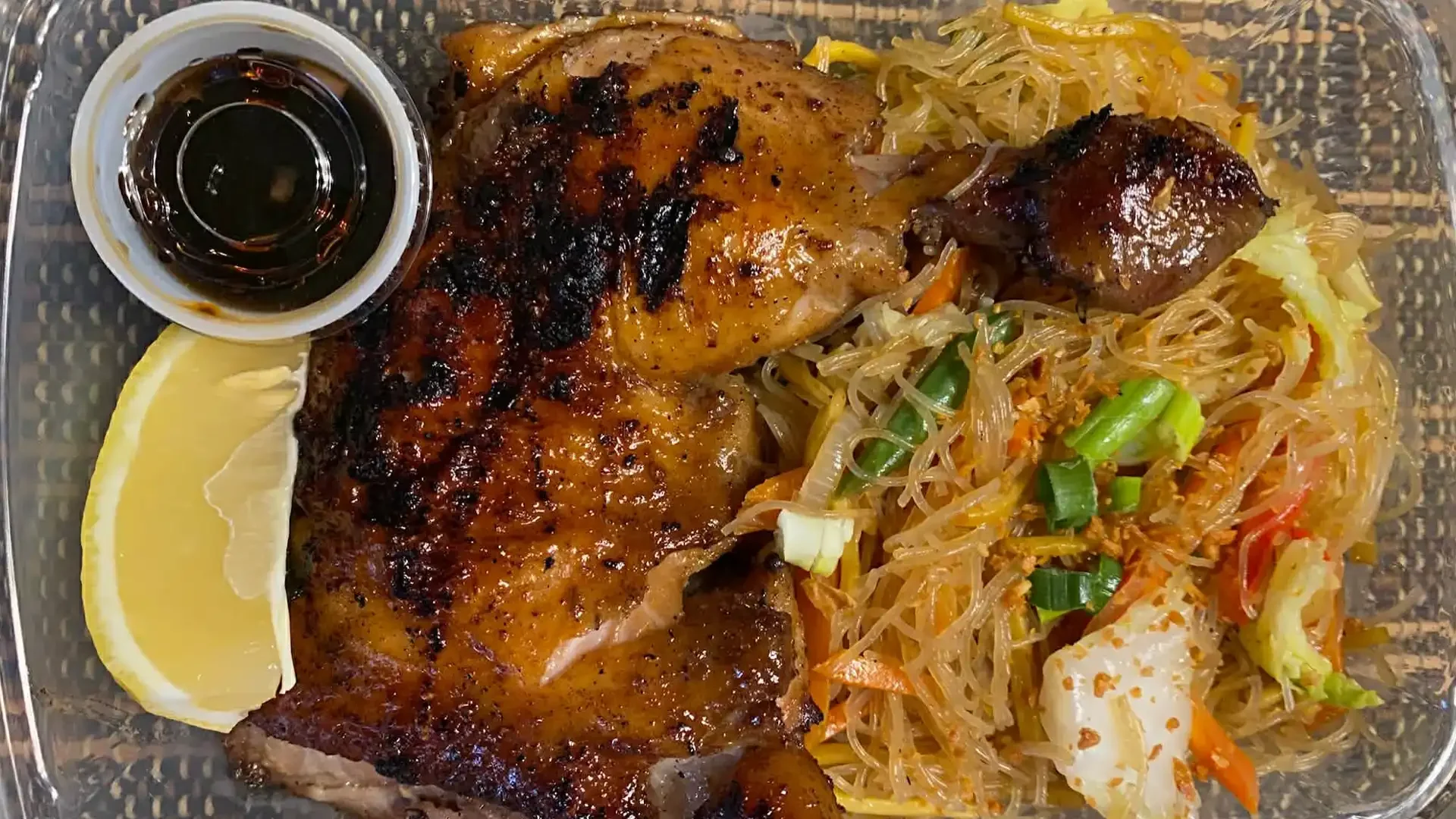 Cooked chicken with a side of Asian-style noodles and vegetables, lemon wedge, and soy sauce in a small container.