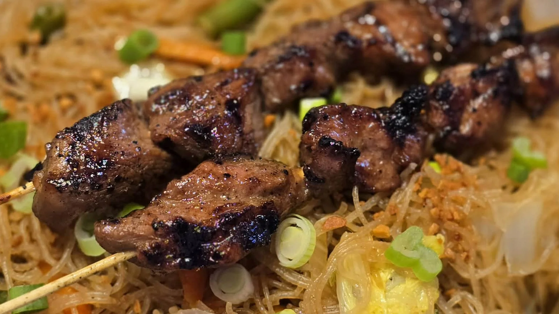 Close-up of grilled beef skewers placed on a bed of stir-fried noodles with chopped green onions and vegetables.