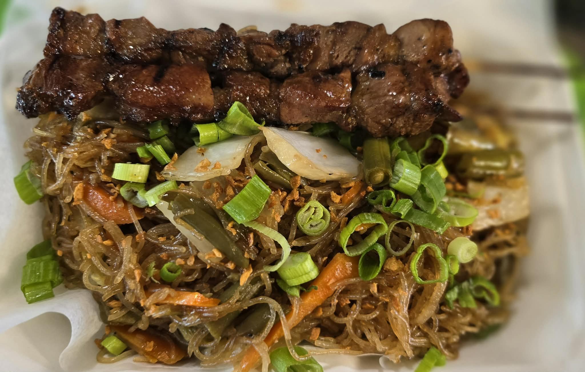 Stir-fried noodles with vegetables topped with grilled meat and chopped green onions.