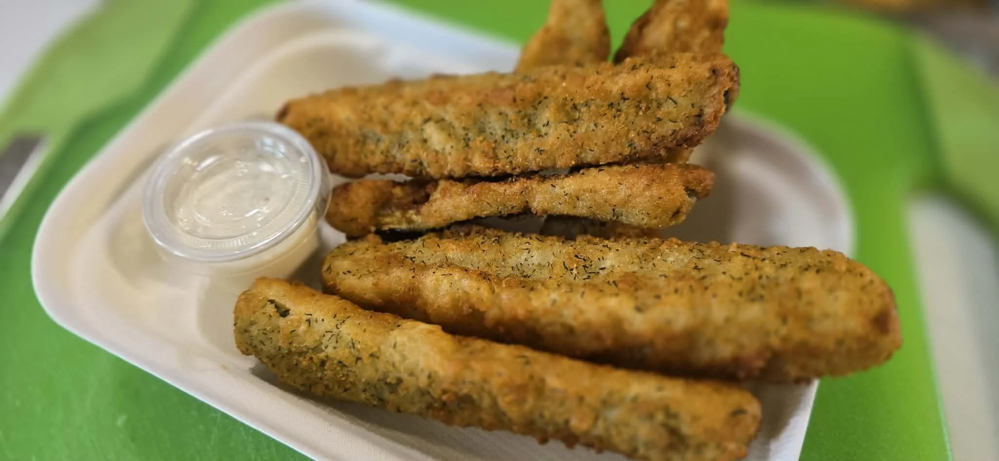 Four breaded fish fillets on a white foam tray with a small container of tartar sauce, on a green table.