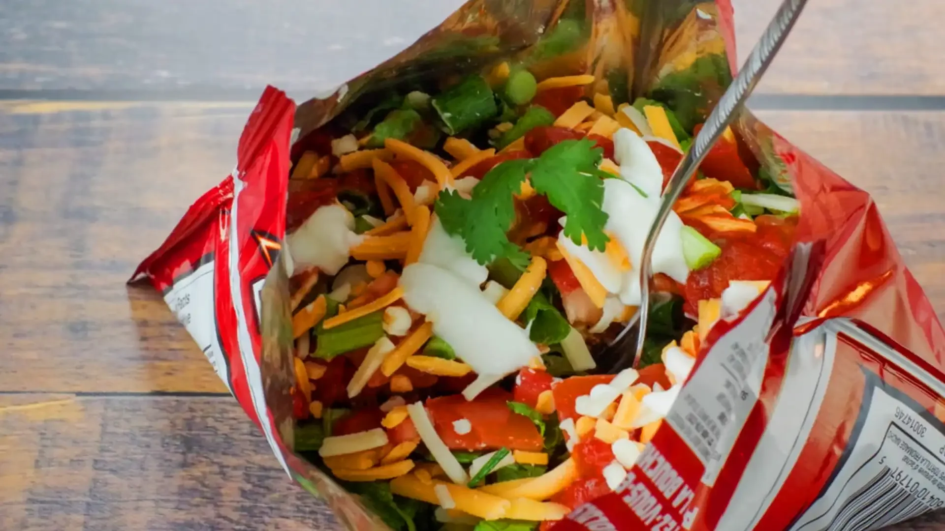Open bag of prepackaged taco salad with shredded cheese, chopped lettuce, tomatoes, and cilantro on a wooden surface.