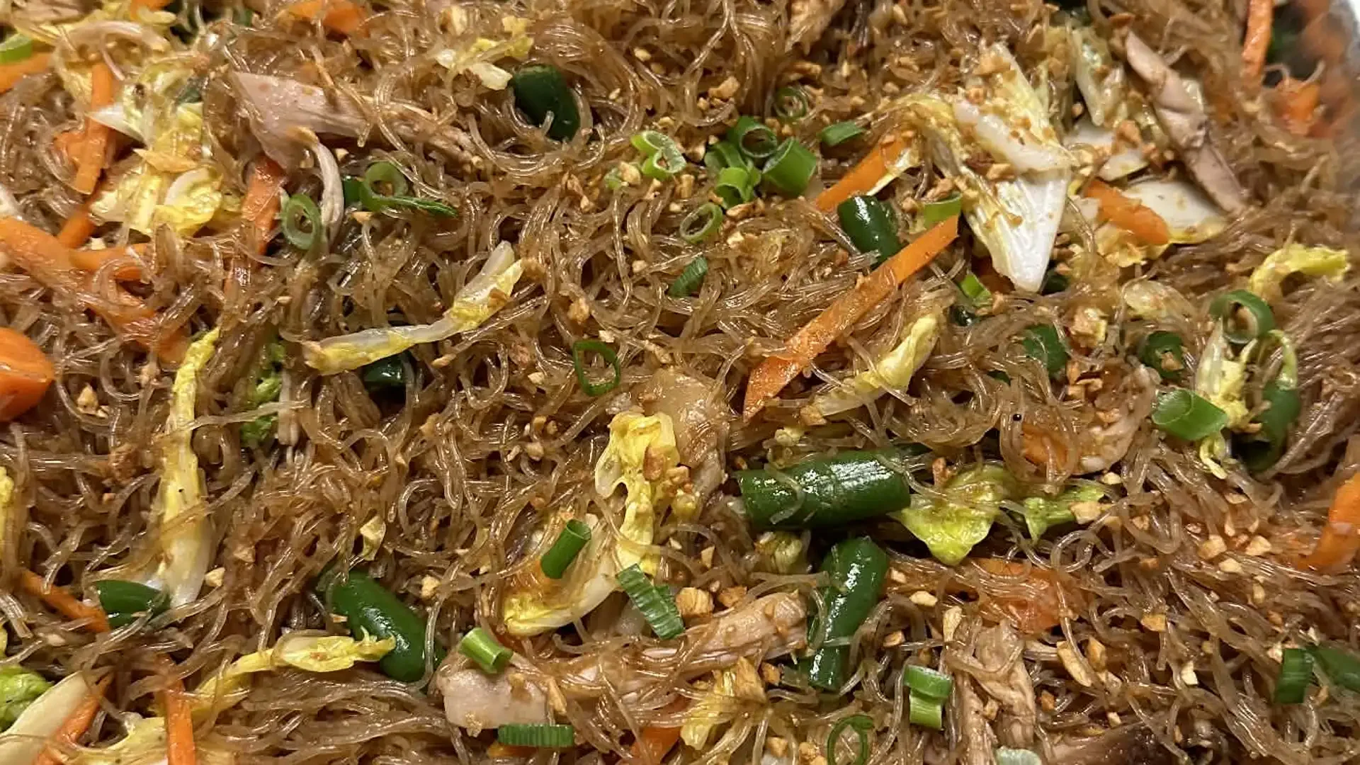 Close-up of a dish with clear glass noodles, chopped vegetables, and green onion garnishing.