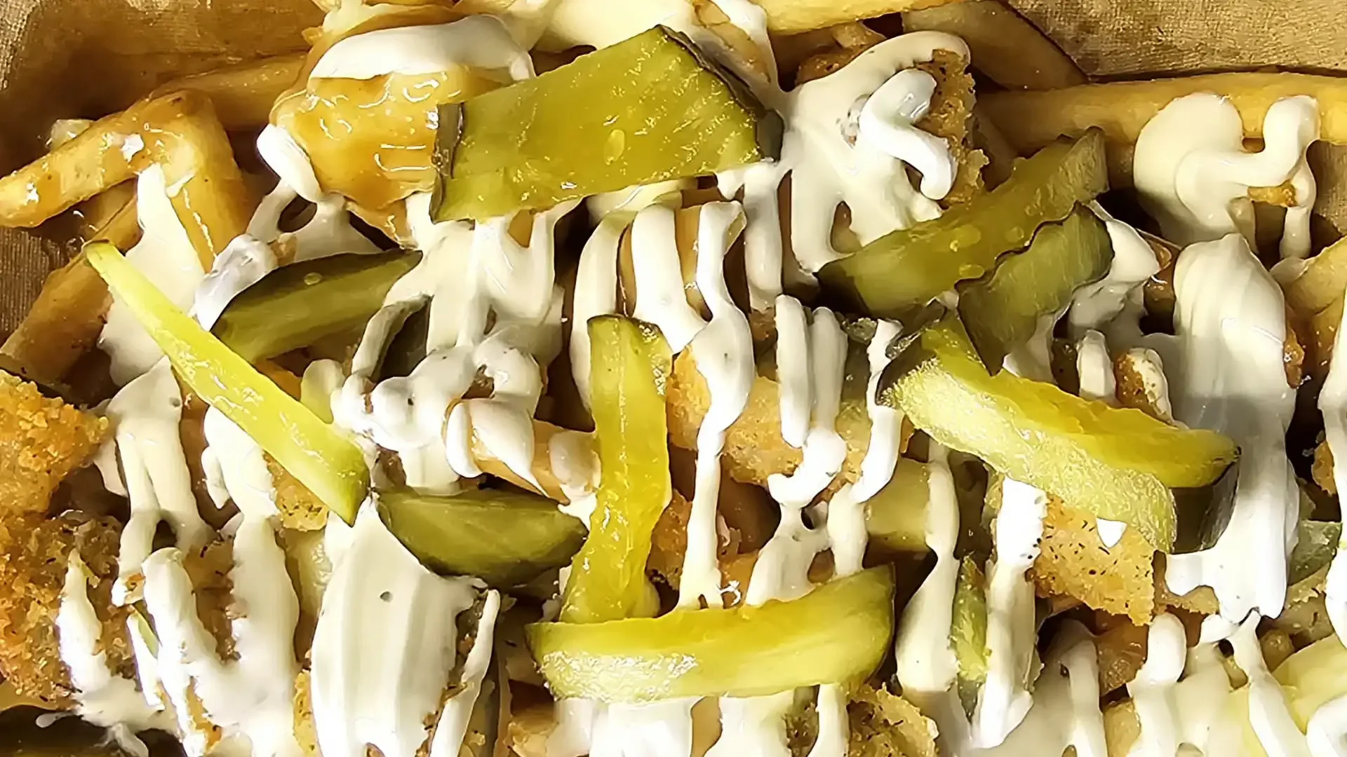 Close-up of a loaded fry basket with pickles, fried chicken pieces, drizzled with white sauce and topped with chopped green peppers.