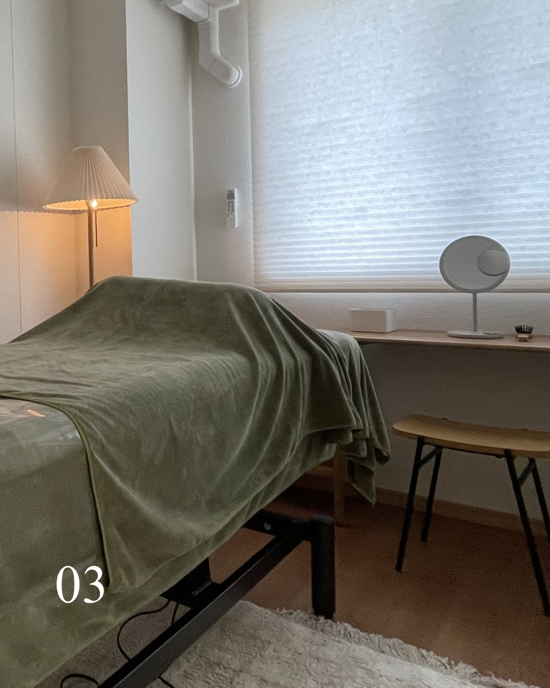 暖色のランプが点灯している部屋の一角。ベッドに緑の毛布がかかっている。窓には白いブラインドが閉まっている。デスクの上には丸い鏡と小さな飾りが置かれている。