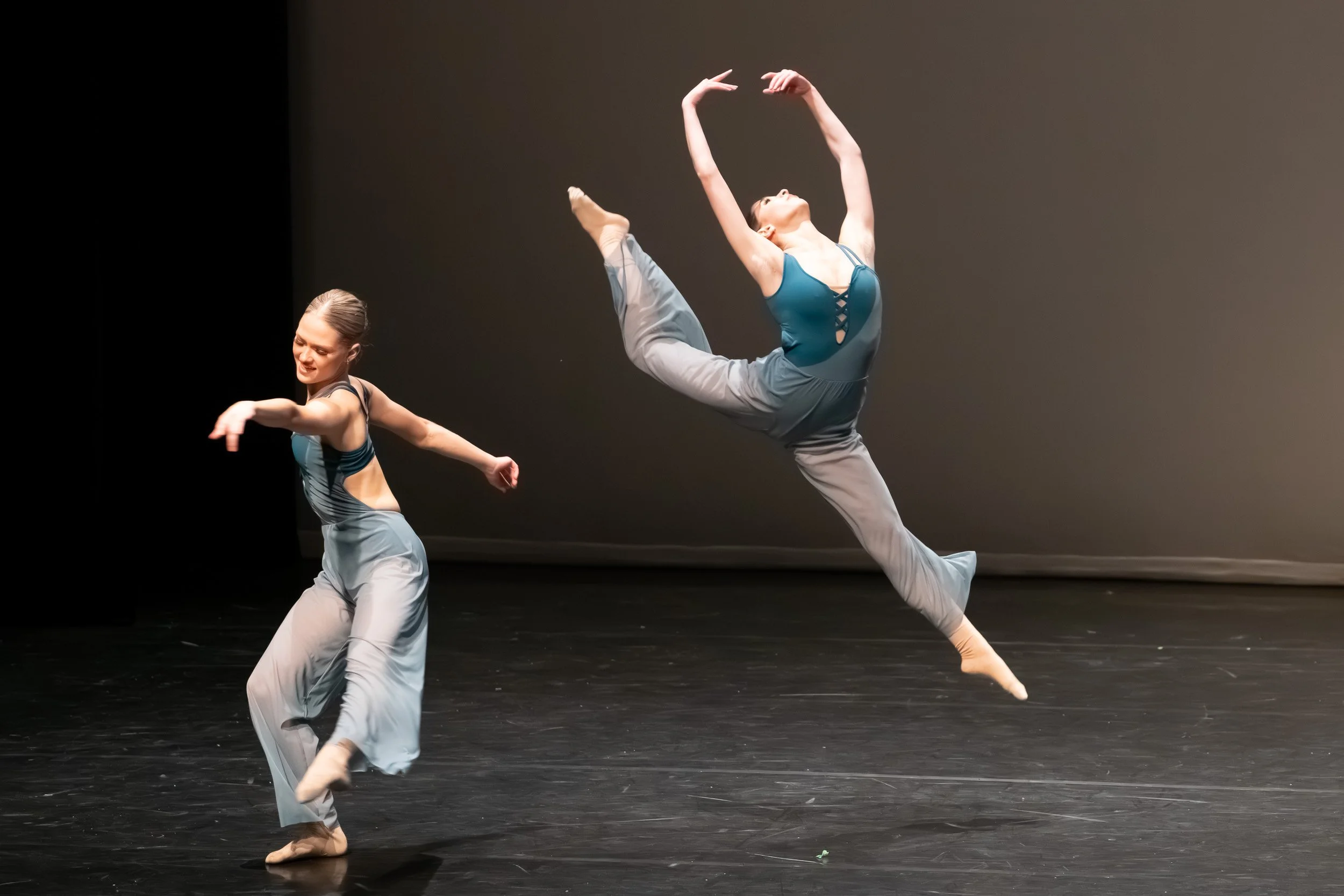 Two female dancers perform on stage, with one leaping mid-air and the other balancing gracefully, both wearing matching blue and gray dance costumes.