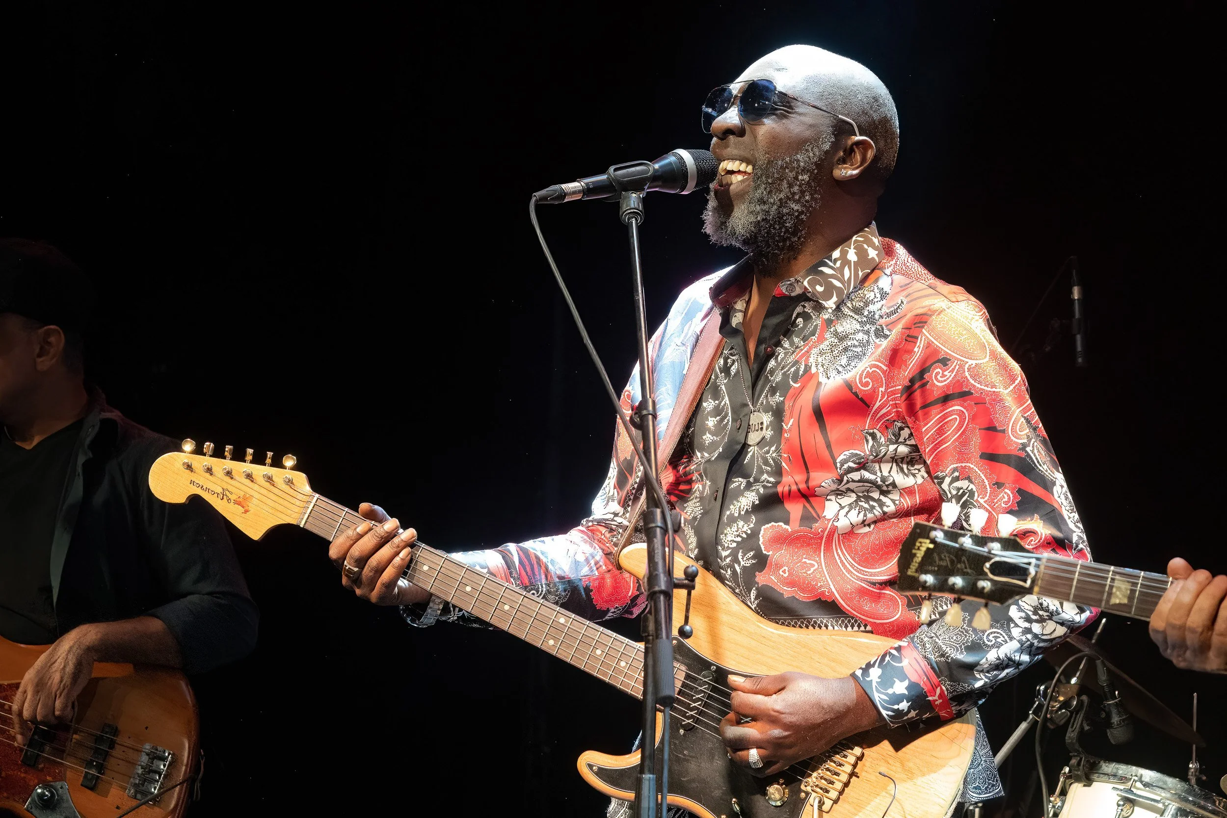 A man with a bald head and a gray beard, wearing sunglasses and a colorful patterned shirt, is singing into a microphone while playing an electric guitar on stage.