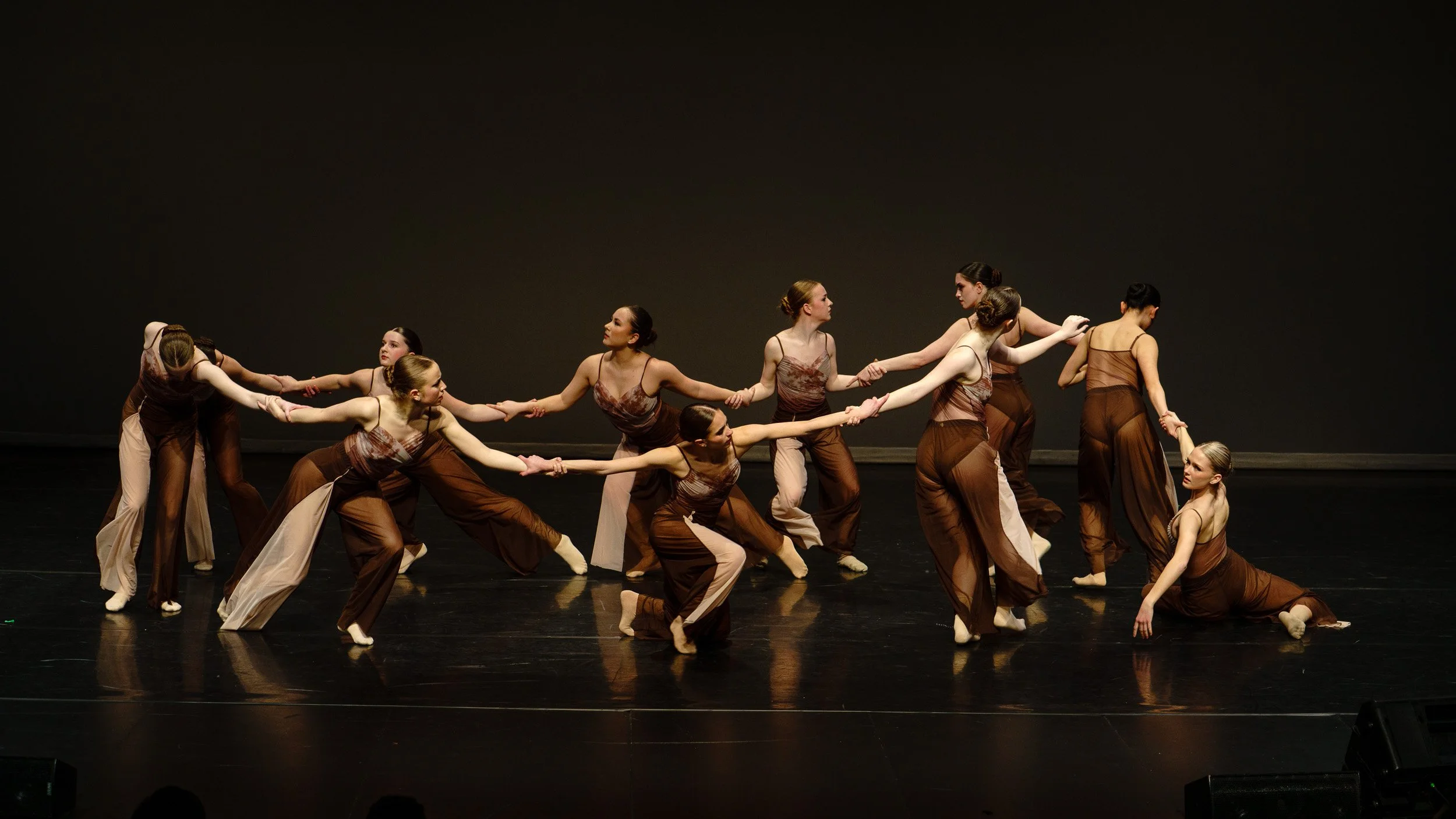 A group of female dancers performing on stage, holding hands and wearing coordinated brown and beige costumes, with some kneeling and others standing.