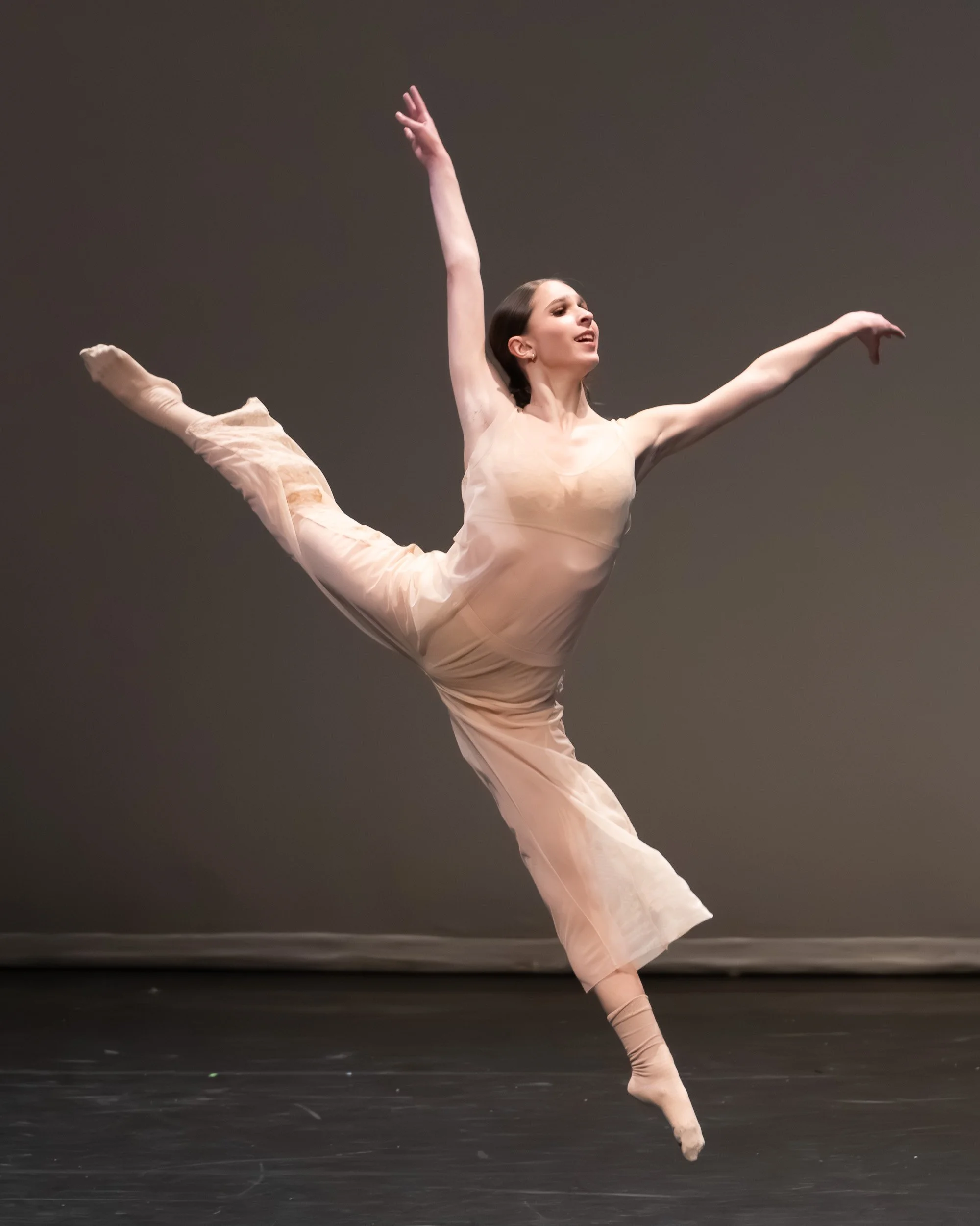A female ballet dancer performing a leap on stage, wearing a beige costume with a neutral background.