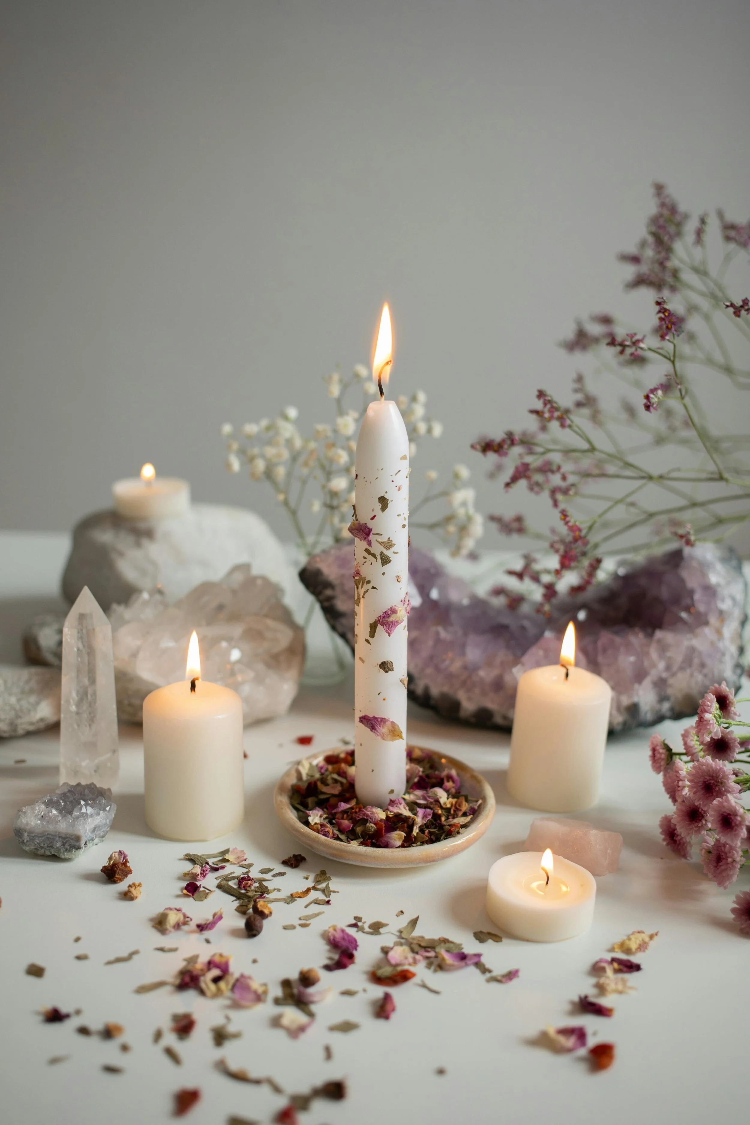 A white candle with dried rose petals burning, surrounded by other lit candles, crystals, and dried flowers on a white surface, with additional crystals and floral arrangements in the background.