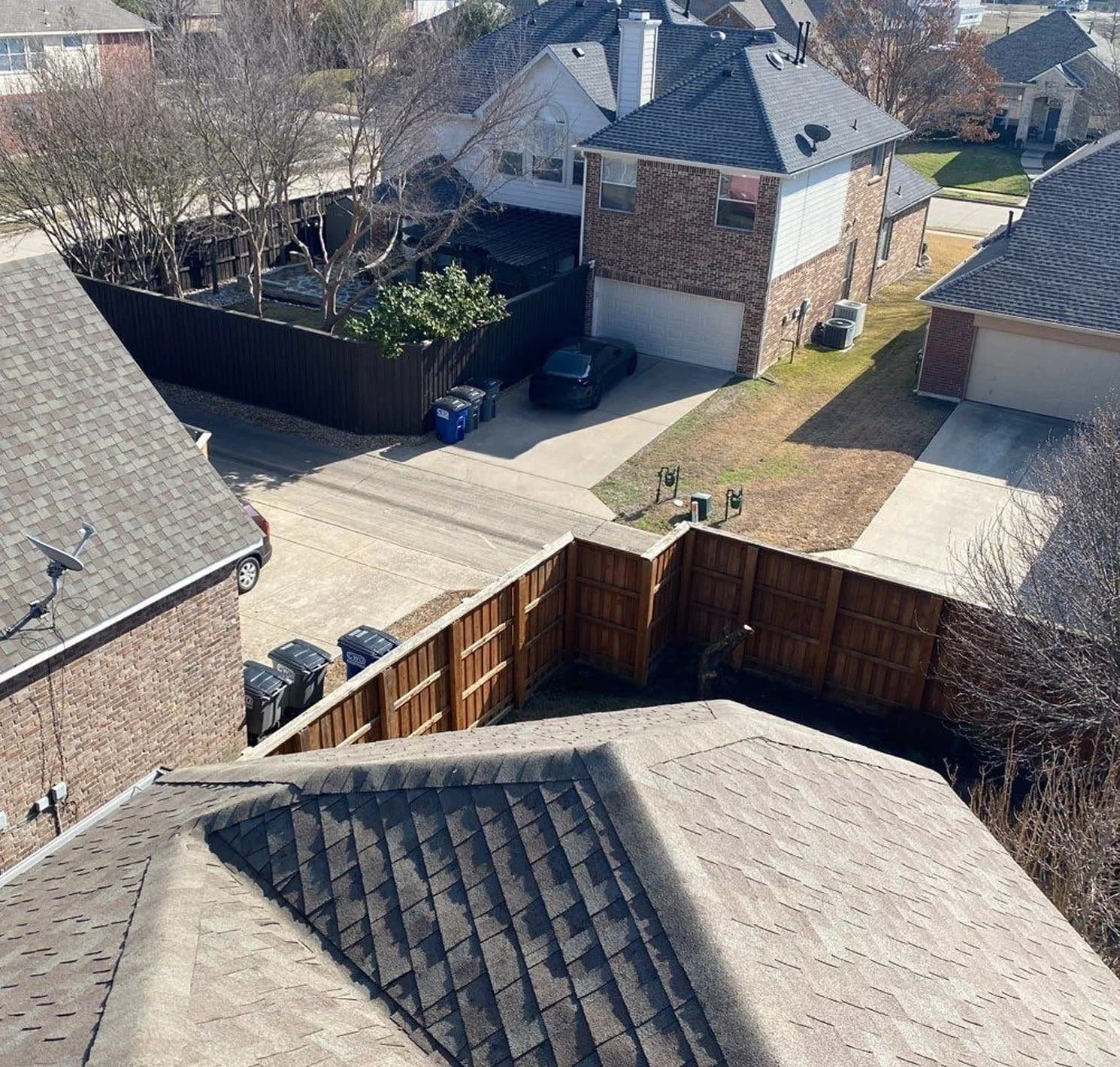 Aerial view of suburban neighborhood with houses, driveways, and yards, including a backyard with a wooden fence and trash bins.