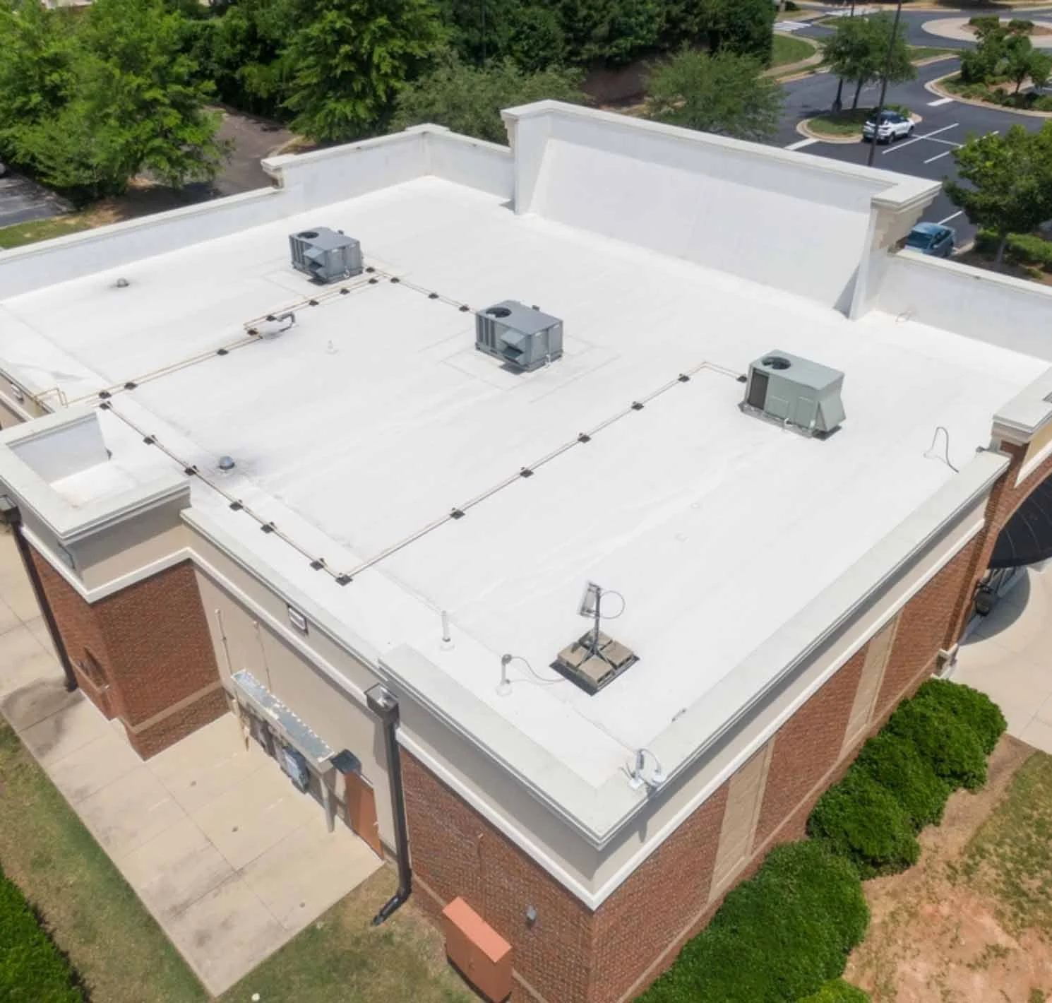 Aerial view of a flat commercial building roof with HVAC units and vents, surrounded by trees and parking lot.