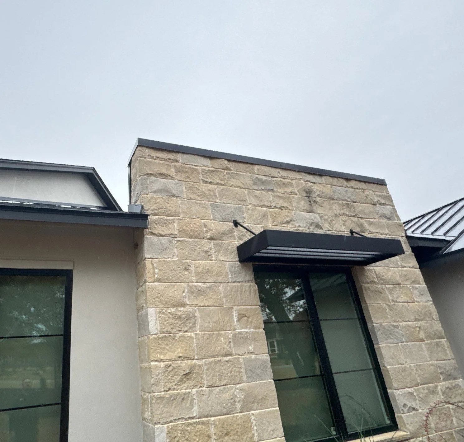Close-up of a modern building's exterior featuring beige stone walls, a large glass window, and a black awning, with a gray overcast sky.