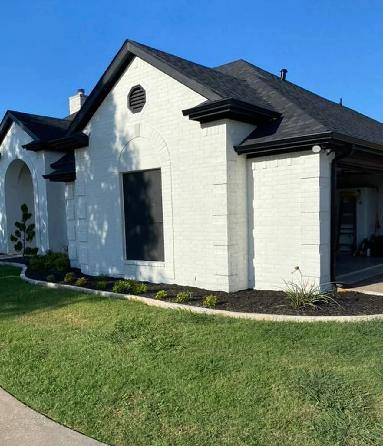 Front view of a white brick house with a gabled black roof, black trim, and a large, arched window covered by a black screen, with a landscaped lawn and sidewalk.