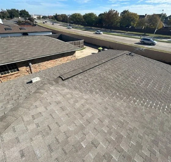 View of a gray shingled rooftop with a chimney showcasing expert roofing services in North Dallas and a street scene in the background showing cars and trees.