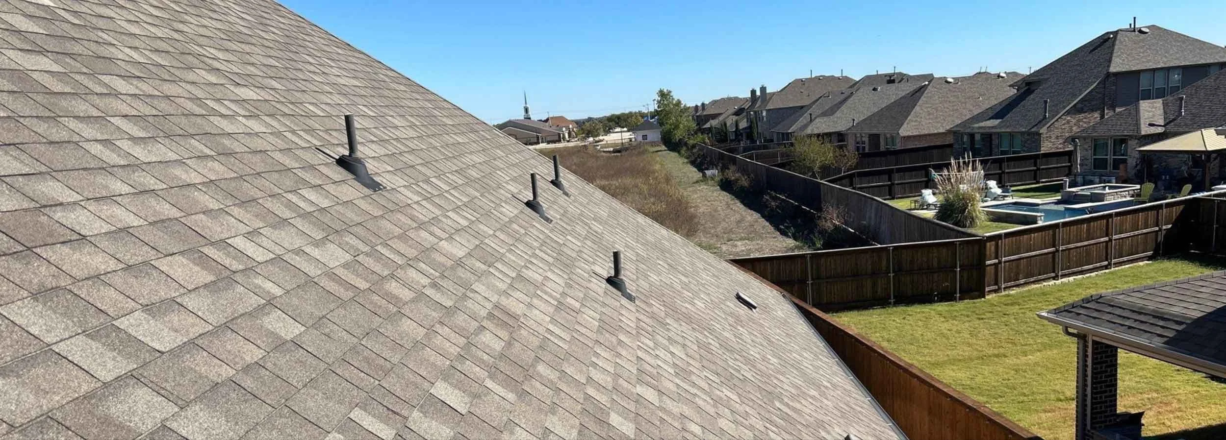 Aerial view of rooftops with asphalt shingles, some roof vents, fenced backyard with a pool and patio, neighboring houses, and a church steeple in the distance under a clear blue sky.