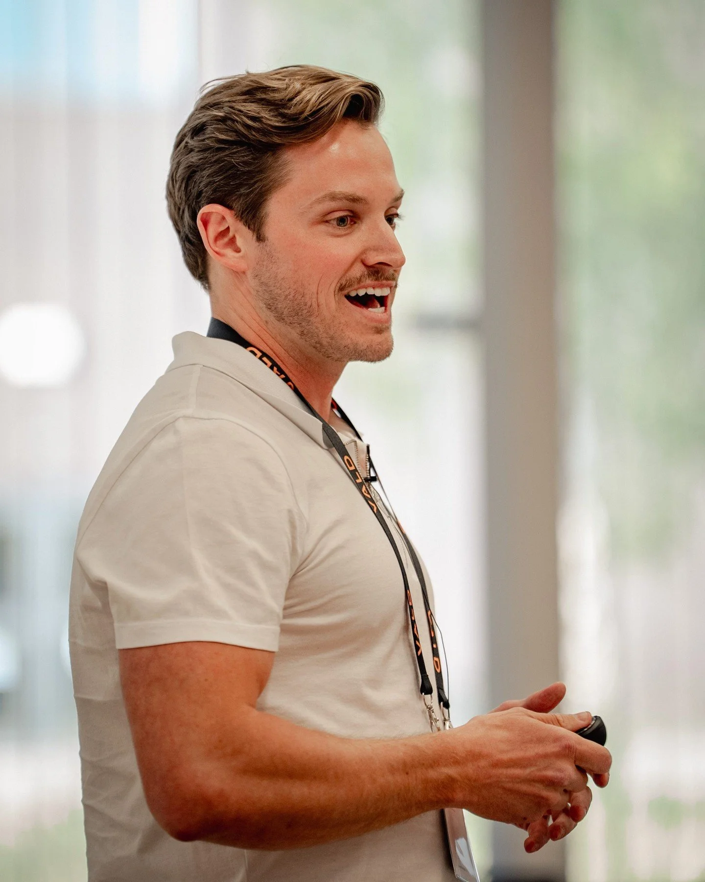 A young man with light brown hair standing indoors, holding a remote control, wearing a beige polo shirt and a lanyard.
