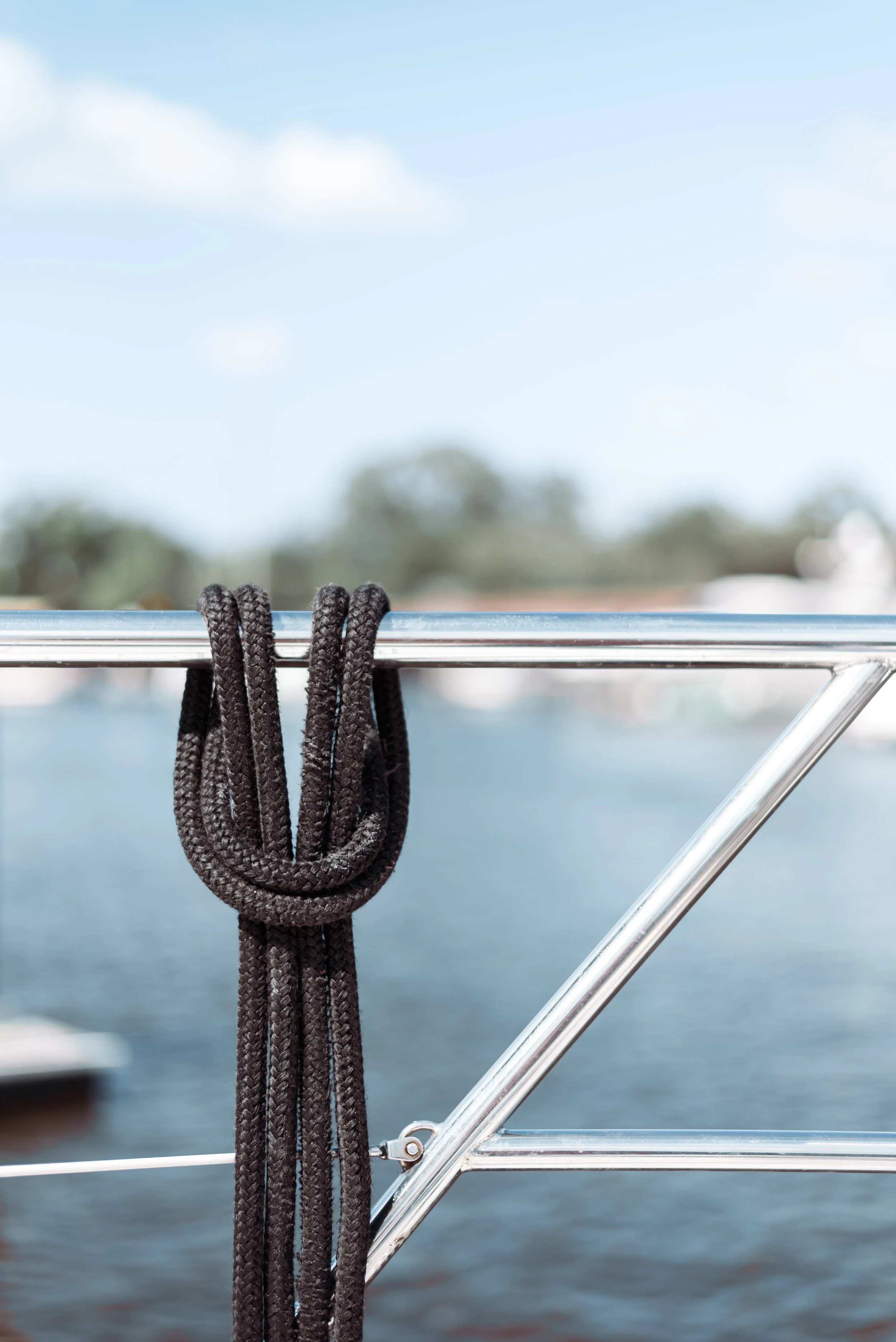 Black coiled rope tied to a metal railing on a boat, with water and blurry trees in the background under a partly cloudy sky.