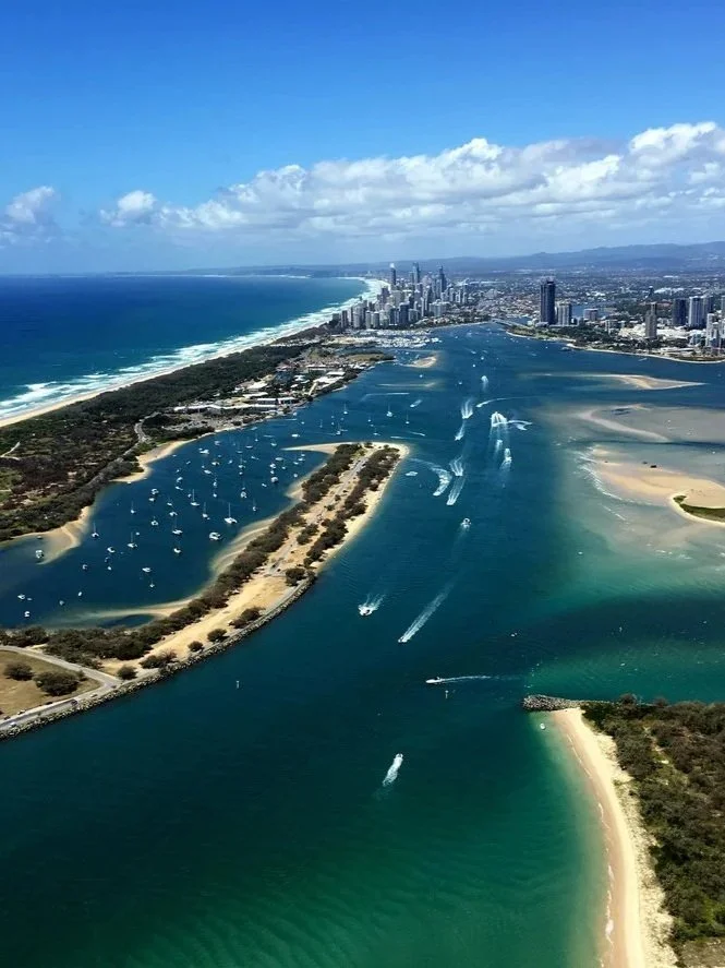 Aerial view of a coastal city with a river running through it, boats sailing on the water, sandy beaches, and a skyline of tall buildings in the background.