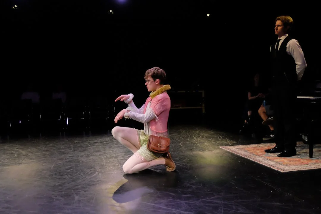 Jack Garrett as Feste in Twelfth Night in colorful, vintage-style clothing kneels on stage while a man in a formal suit stands nearby, with a dark background and stage lights.