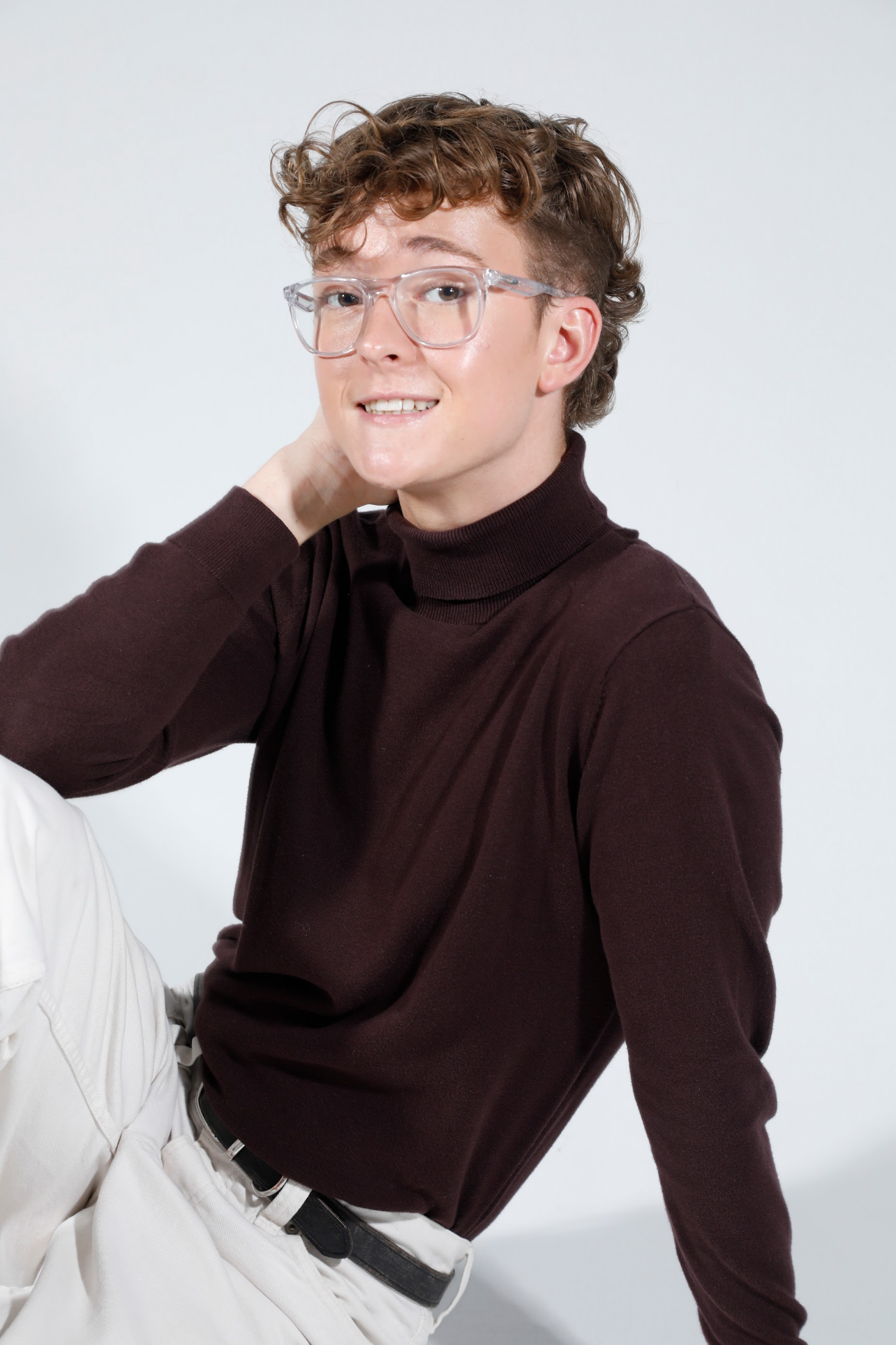 Jack Garrett, a young person with curly hair and glasses wearing a dark turtleneck sweater and white pants, posing against a plain white background.