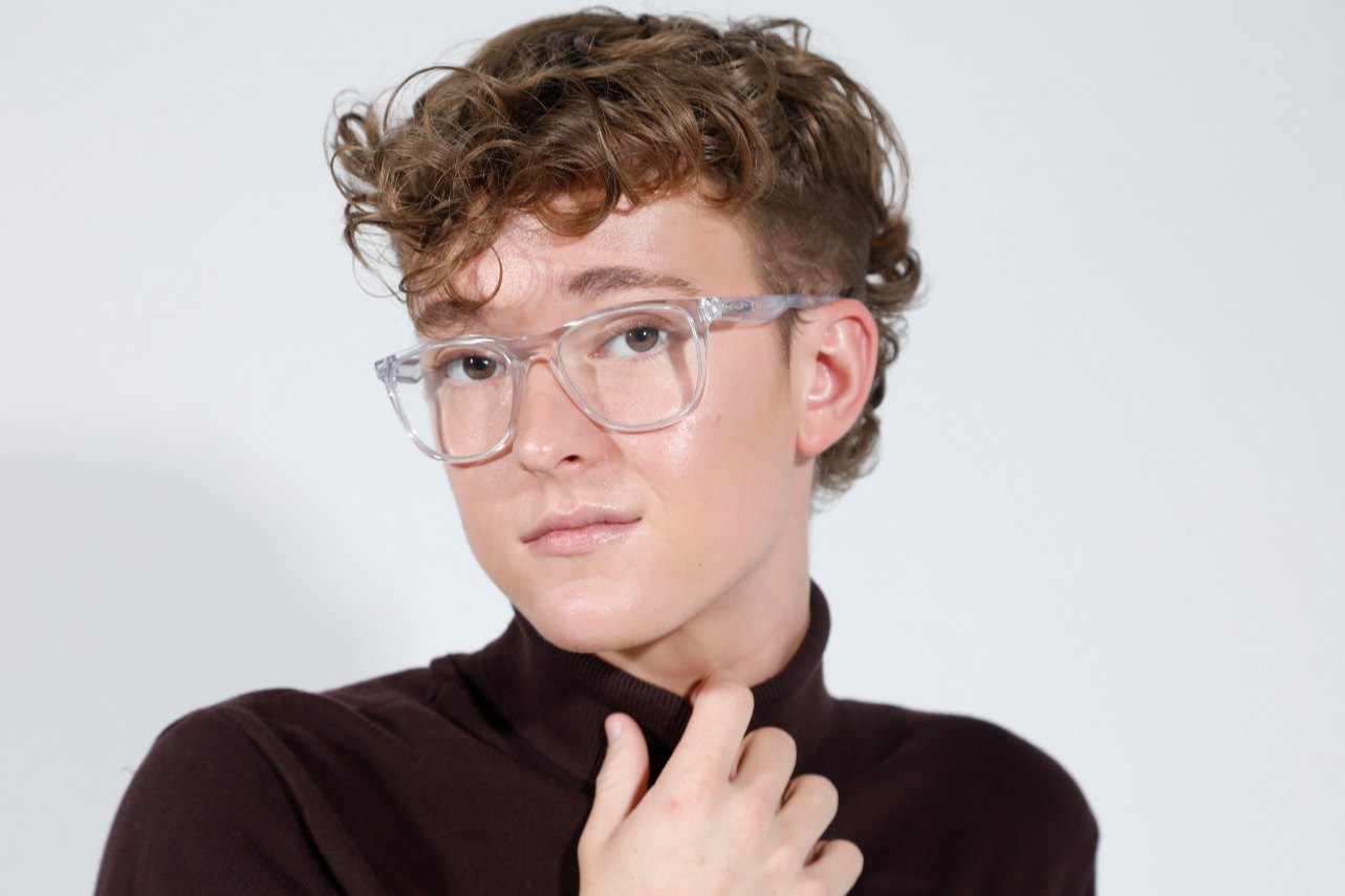 Jack Garrett, a person with short curly hair wearing large clear glasses and a dark turtleneck, touching their neck, against a plain white background.