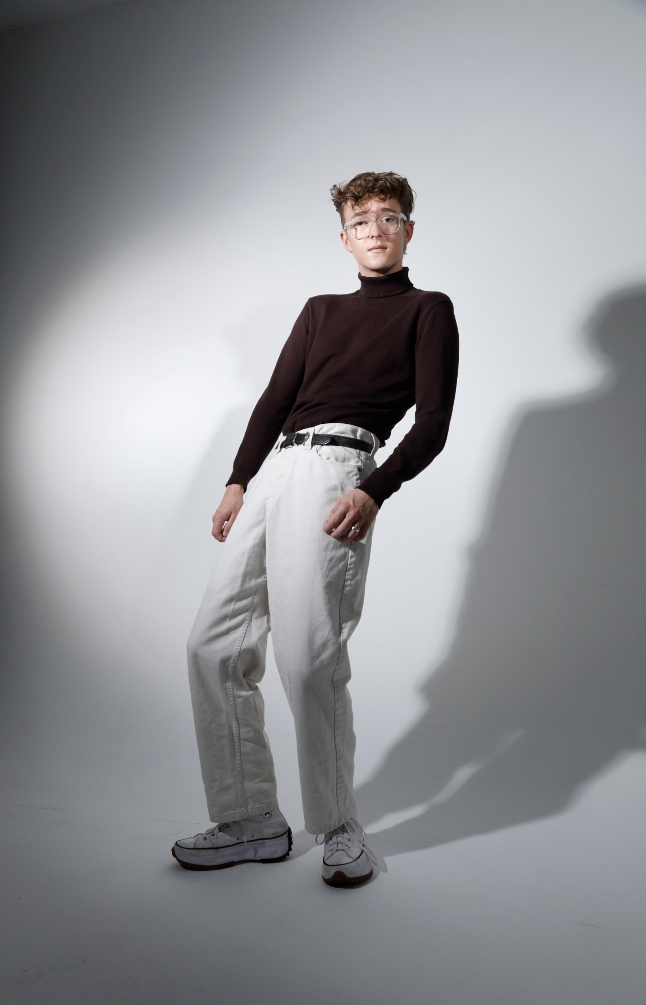 Jack Garrett, a person with short curly hair, glasses, black turtleneck sweater, white baggy pants, and sneakers standing in a photo studio with a soft shadow on a plain background.