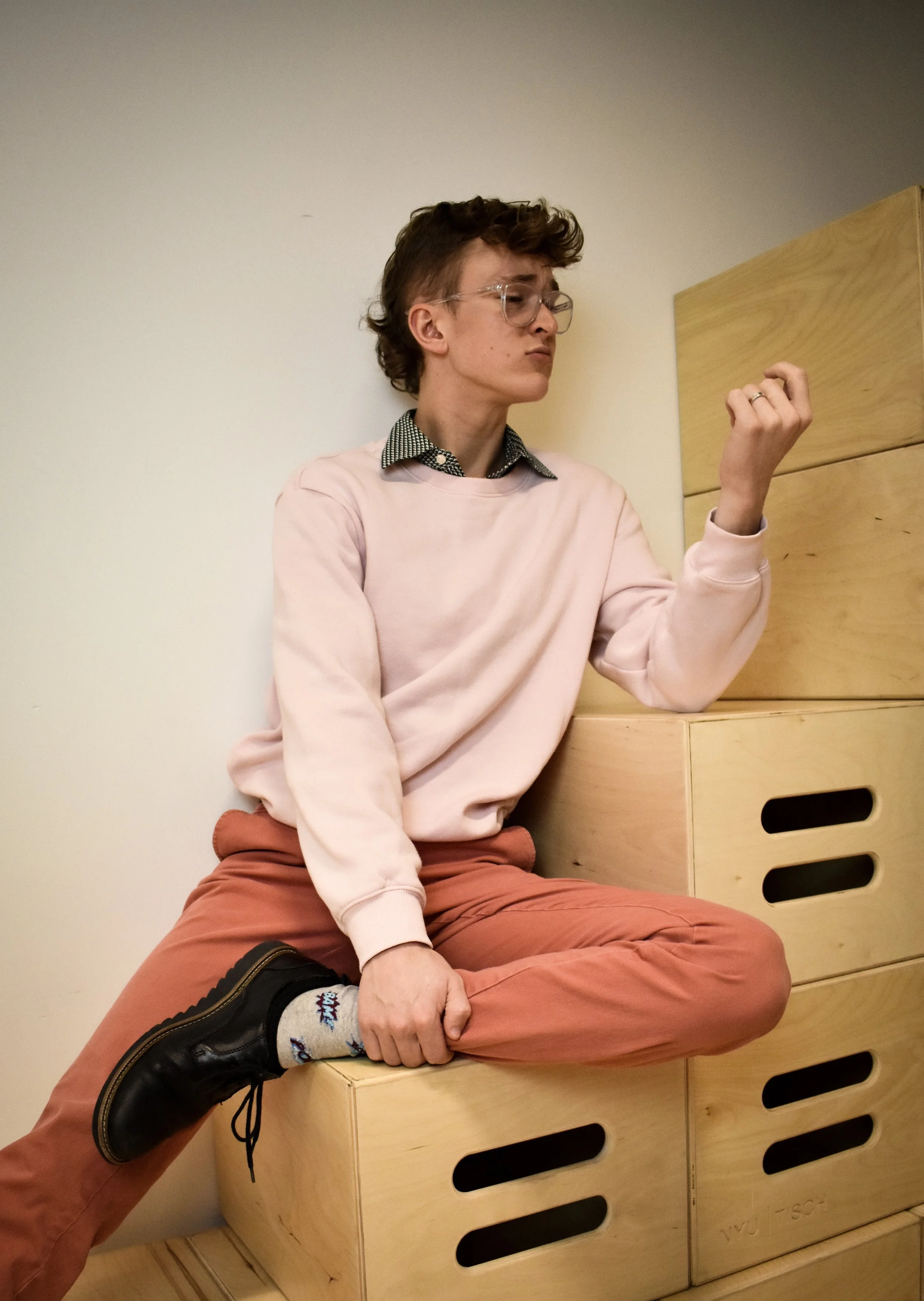 Jack Garrett as Green in Richard II, sitting on wooden boxes, wearing glasses, pink sweatshirt, patterned shirt underneath, and brown pants, with one leg crossed over the other and looking at their fist.