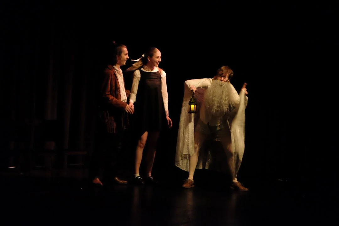 Jack Garrett as Feste with a long beard performing on a dimly lit stage, with black curtains in the background. The person with the beard holds a lantern and is wearing a robe.