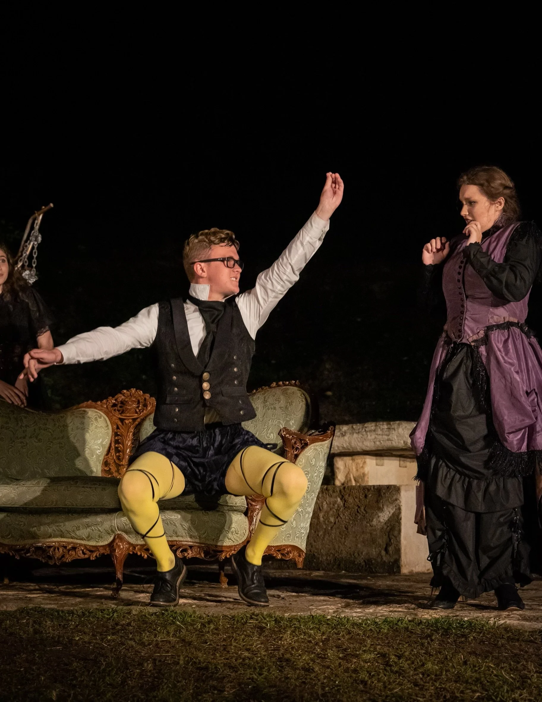 Jack Garrett as Malvolio in Twelfth Night with yellow tights and black shoes, sitting on an ornate sofa, raising his hand. A woman in period costume stands nearby, appearing surprised or concerned. The scene is set outdoors at night.