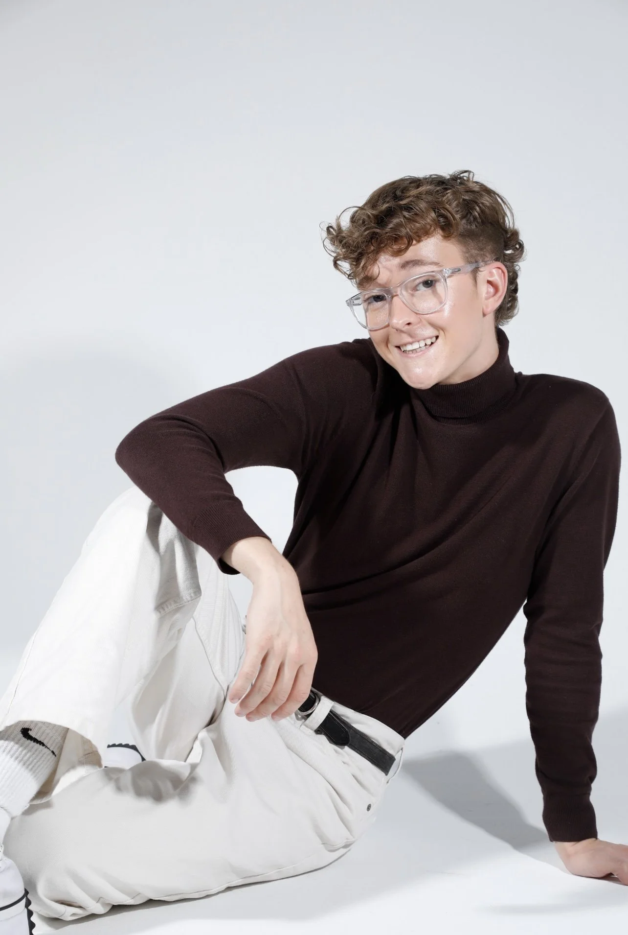 Jack Garrett, a young man with curly hair and glasses, sitting on a white surface, smiling, wearing a black turtleneck and white pants against a plain light background.
