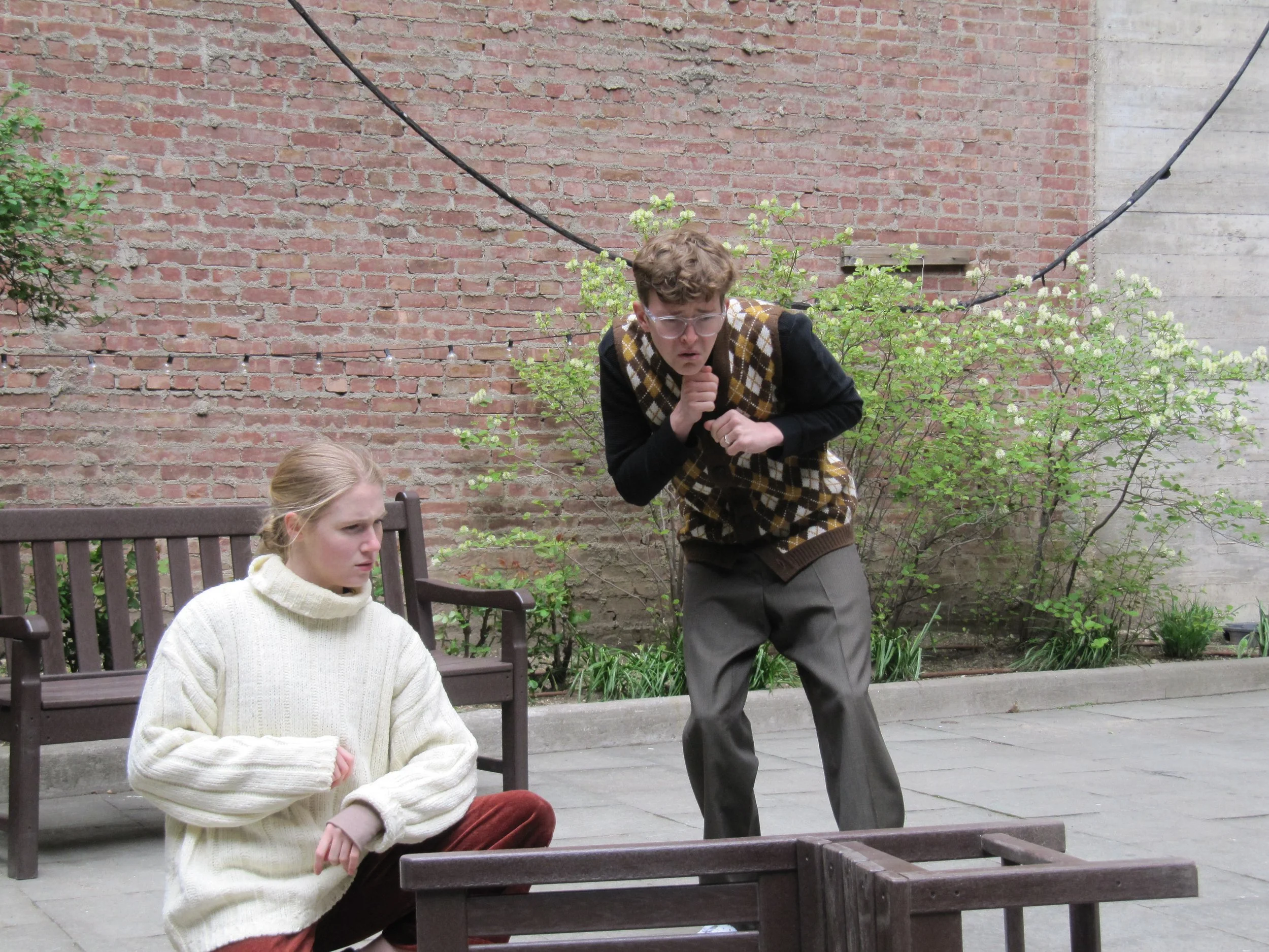 Jack Garrett as Dull in Love's Labour's Lost, looking at something on the ground with a worried or surprised expression, in an outdoor setting with brick walls and green plants.