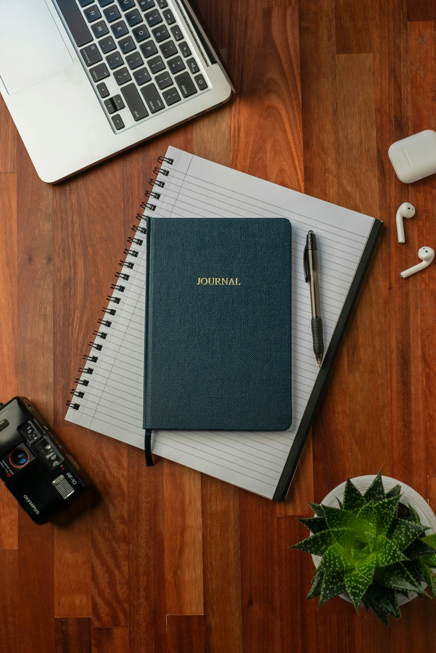 A flat lay of a workspace on a wooden desk, featuring a silver laptop, a black ballpoint pen, a blue journal, spiral notebooks, edible AirPods case, a camera, and a potted green succulent plant.