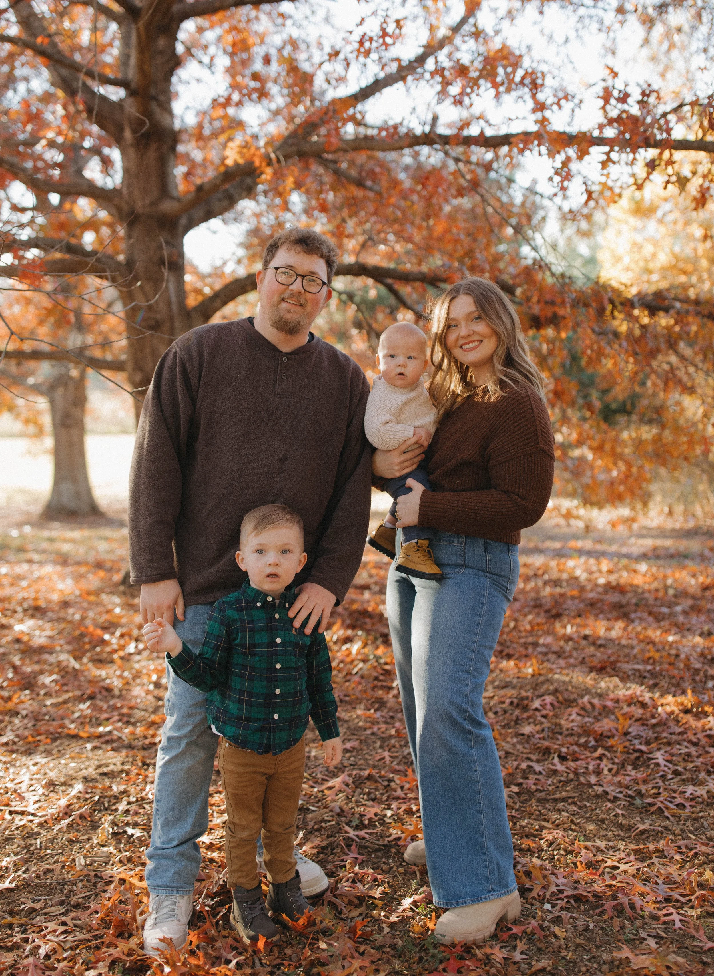 Family of four standing outdoors on a fall day with orange and brown leaves, a tree in the background, and sunlight filtering through the branches.