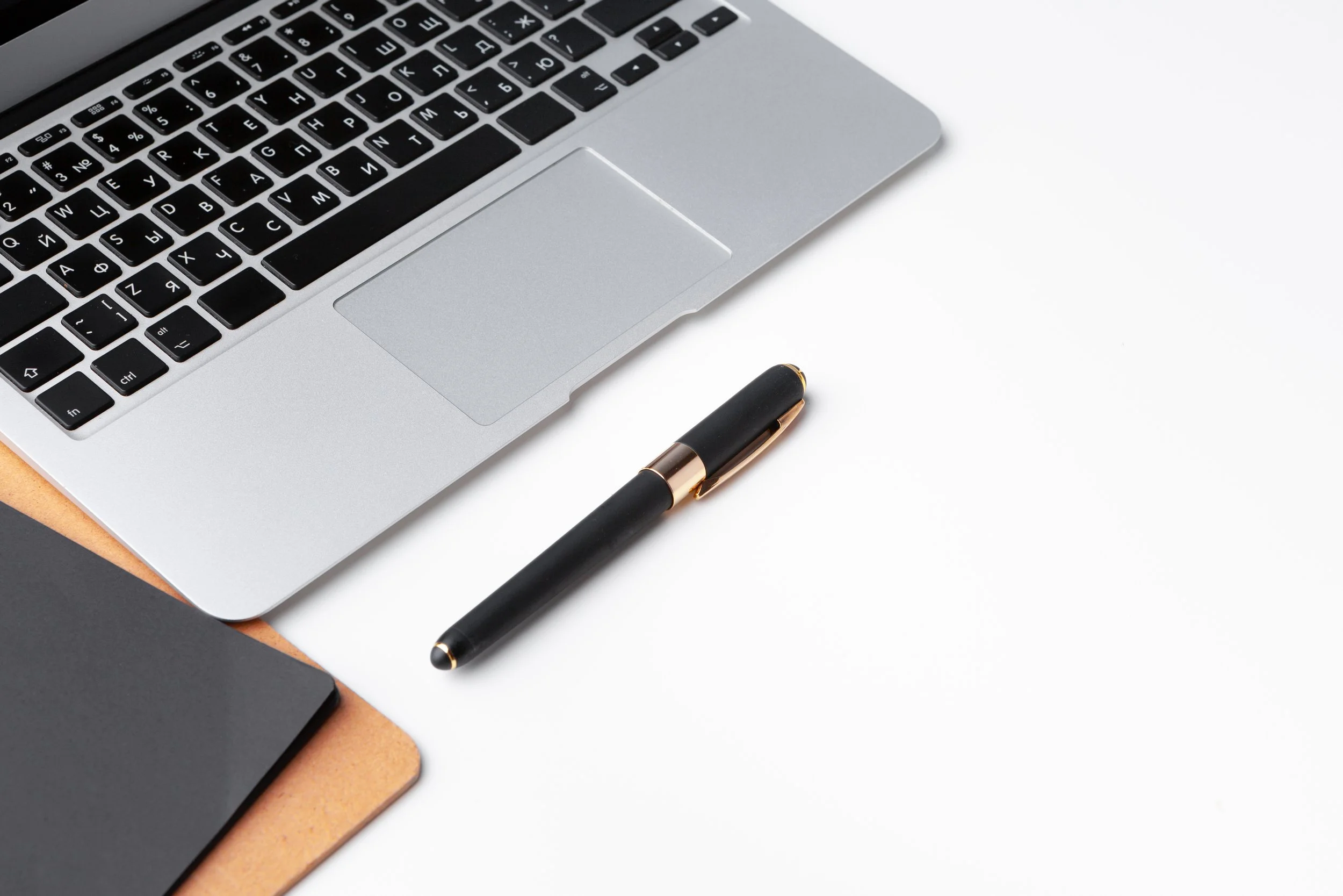 A partial view of a silver laptop keyboard, a black and gold pen, a black notebook, and a cork clipboard on a white desk surface.
