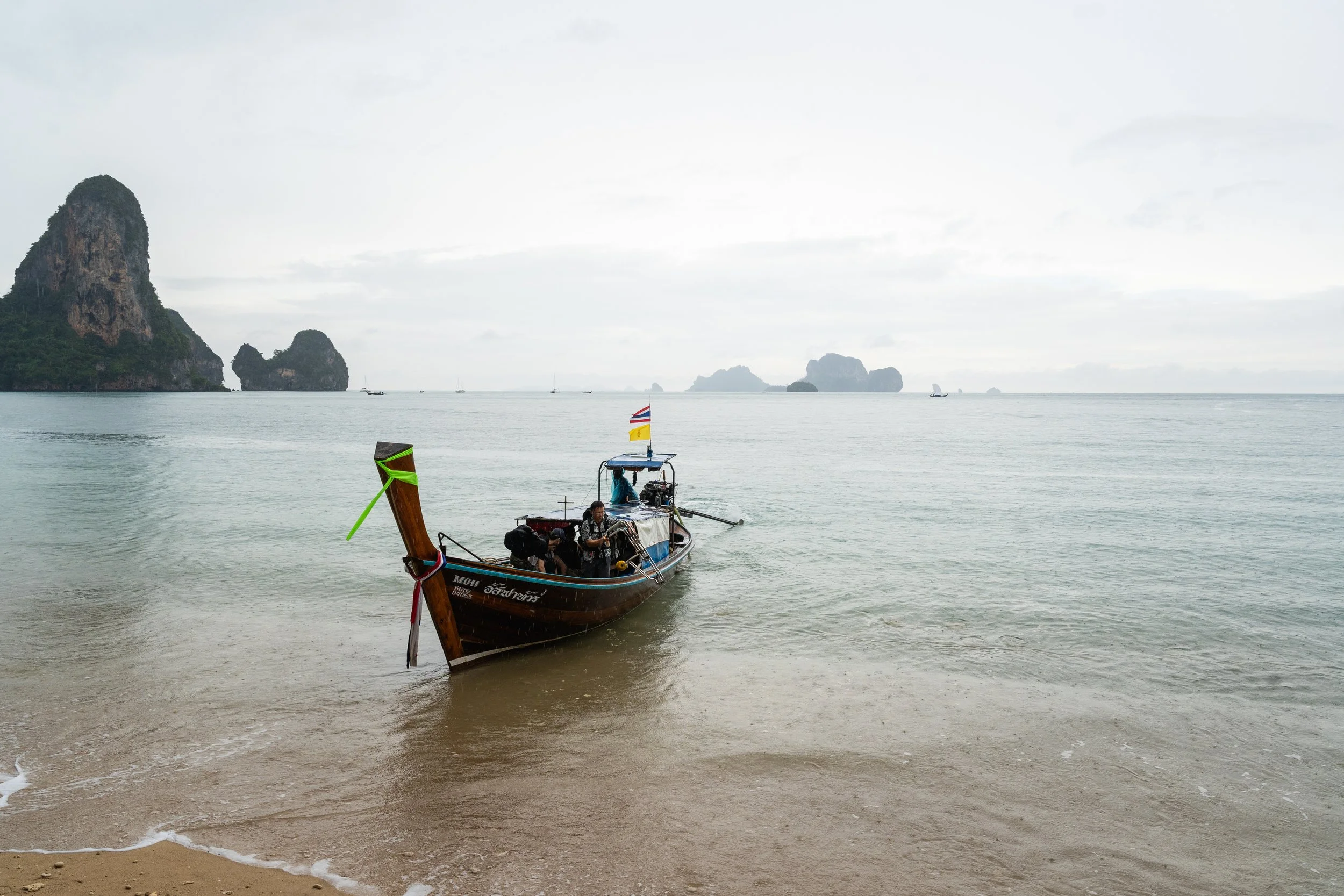 A small boat with several people onboard is anchored near a sandy beach on a cloudy day, with distant islands and rock formations visible across the water.