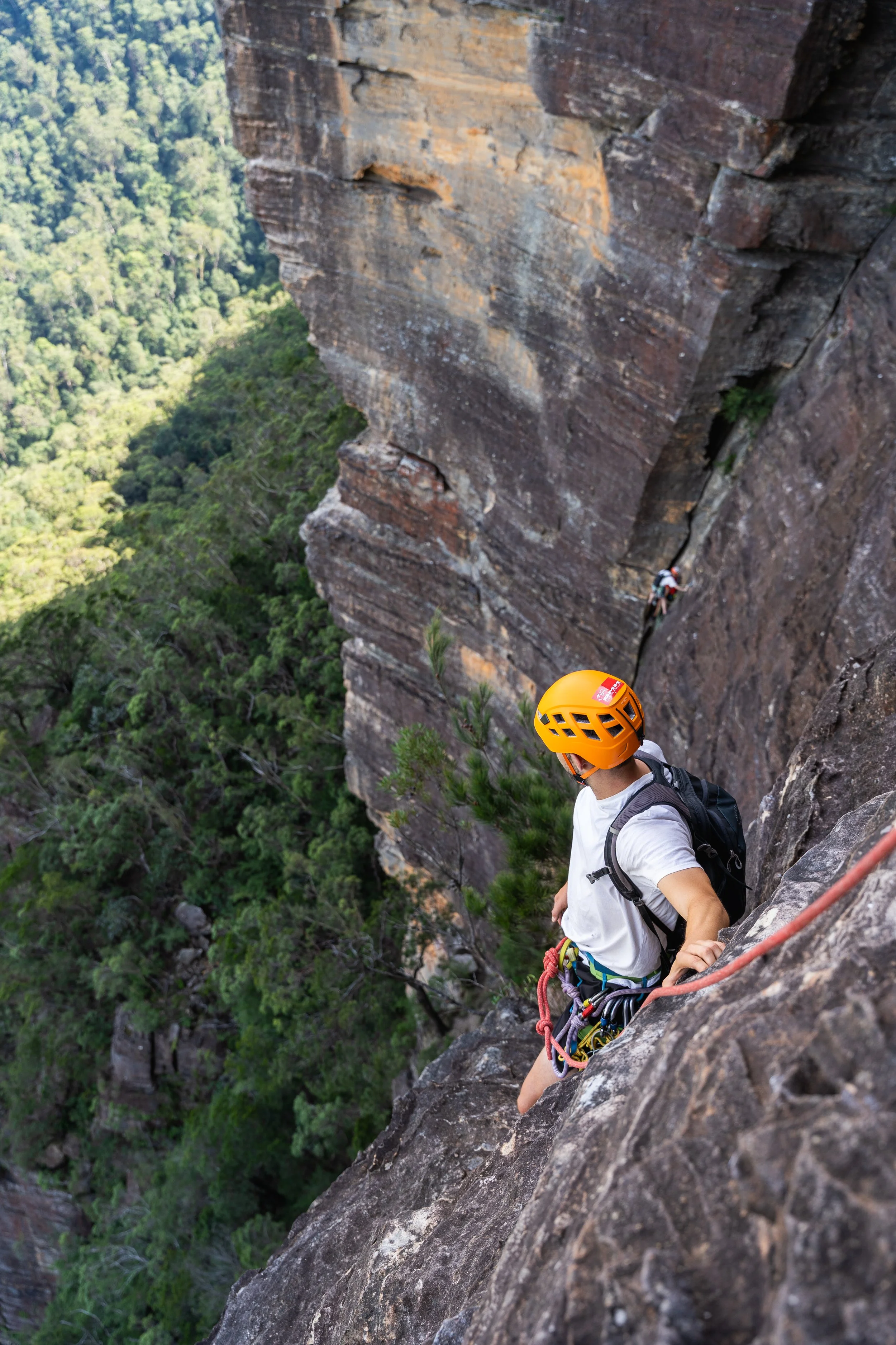 A rock climber wearing an orange helmet, white t-shirt, and climbing gear is ascending a steep cliff face, with another climber visible higher up on the rock.