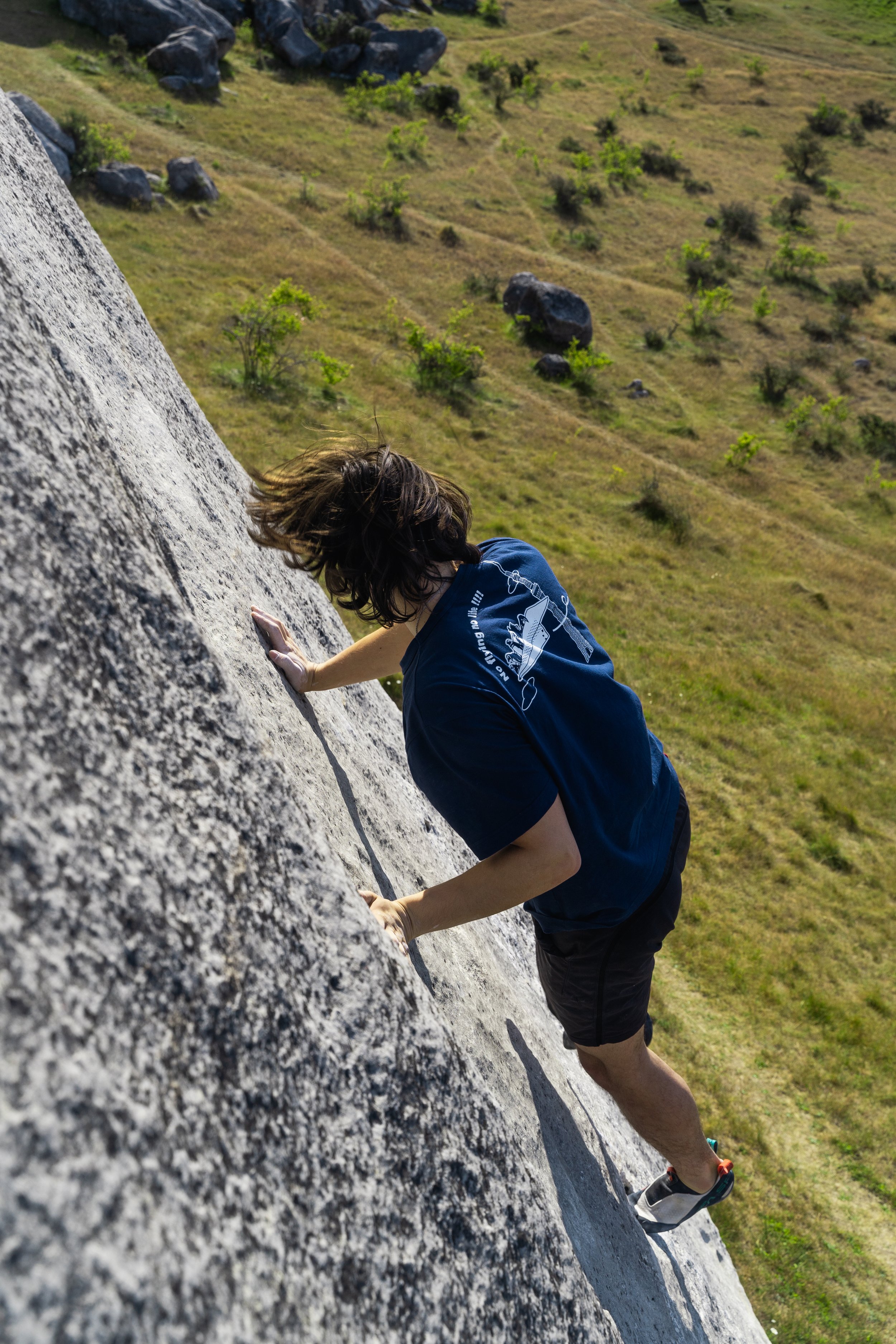 Person rock climbing on a steep outdoor rock face with grassy landscape and rocks in the background.