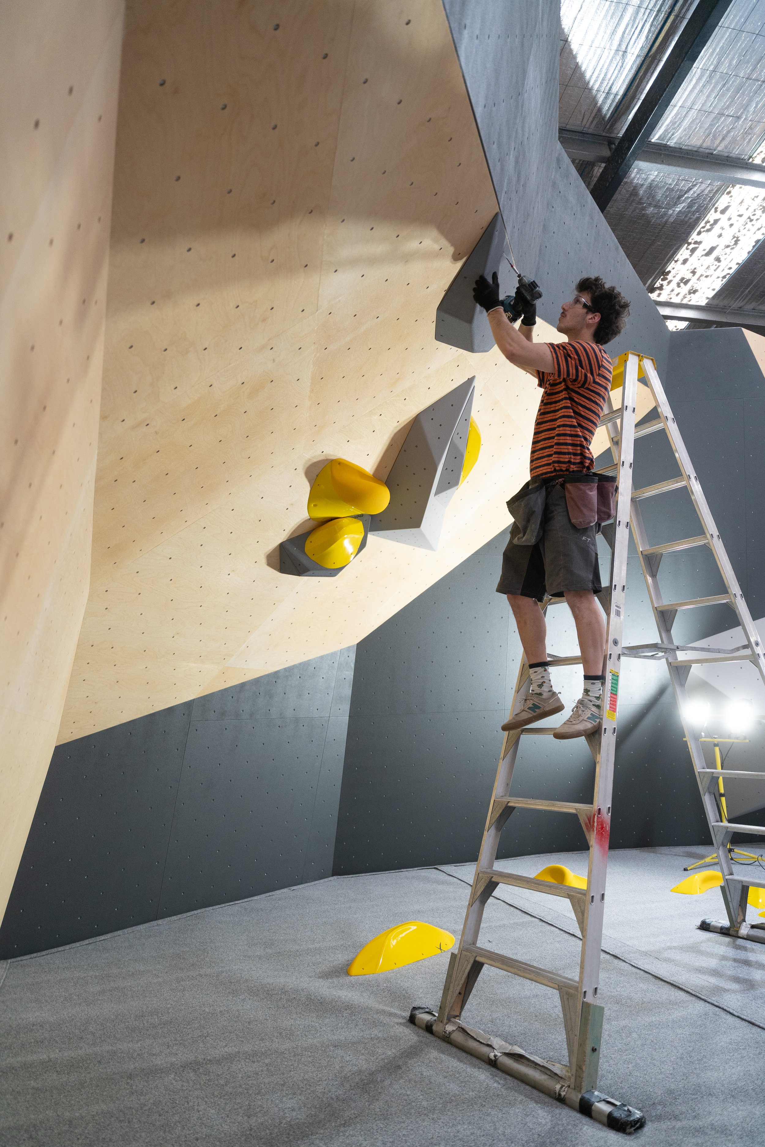 A man standing on a ladder installing holds on an indoor climbing wall. The climbing wall has a wooden section with various rock climbing holds, including yellow and gray ones.