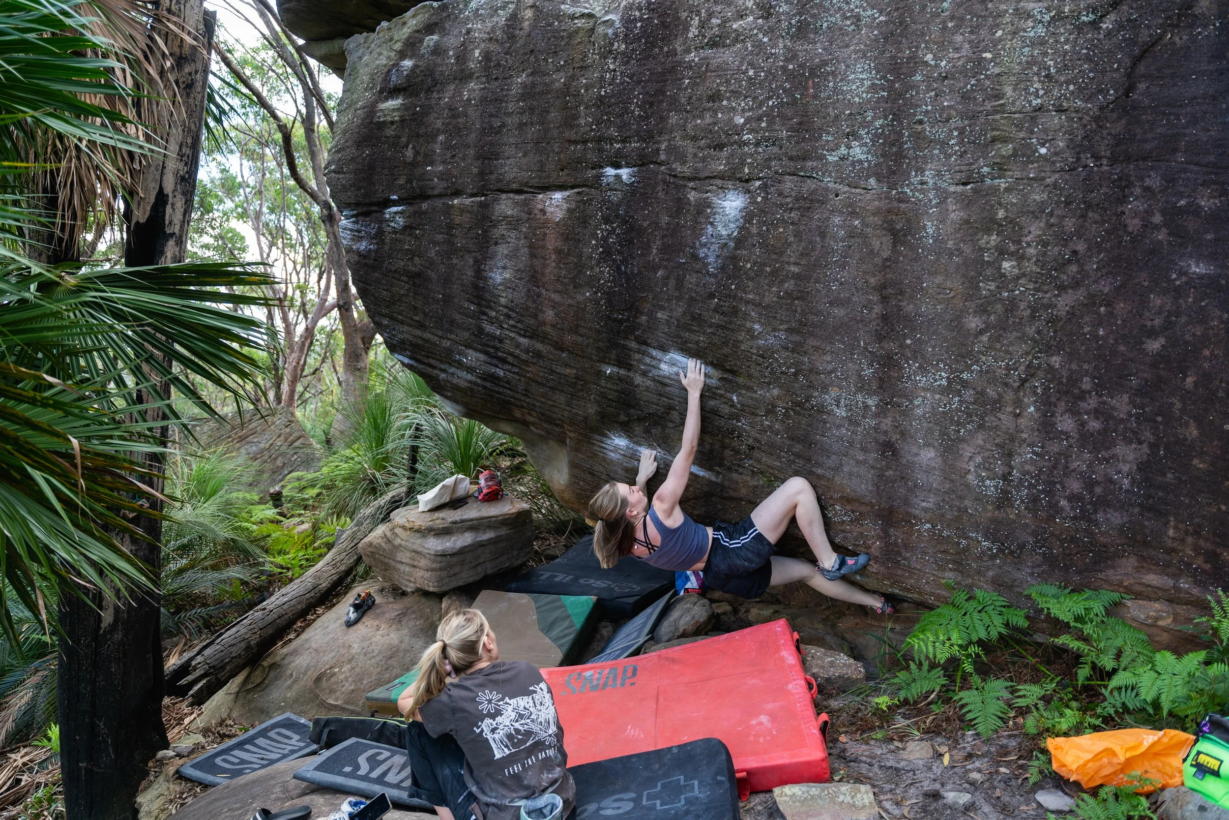 A woman climbing a large overhanging boulder in a forested area, with two women sitting on mats watching. One woman is reaching up to grip the rock while the others prepare gear.