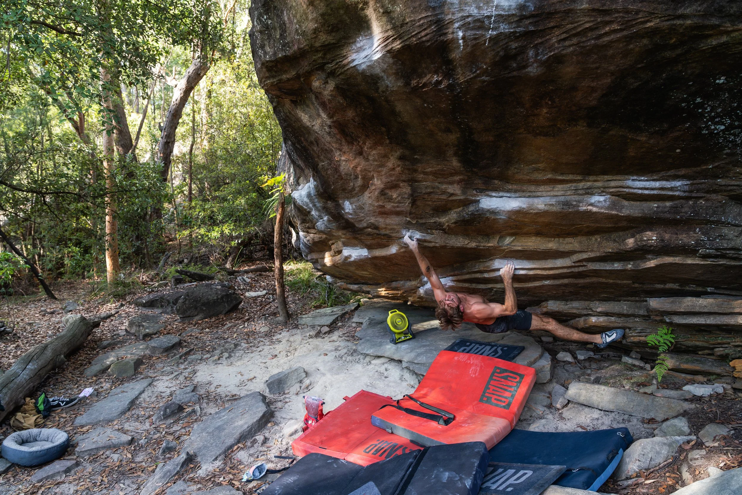 A man bouldering outdoors under a large overhanging rock, with crash pads, a chalk bag, and climbing equipment on the ground surrounded by trees and foliage.