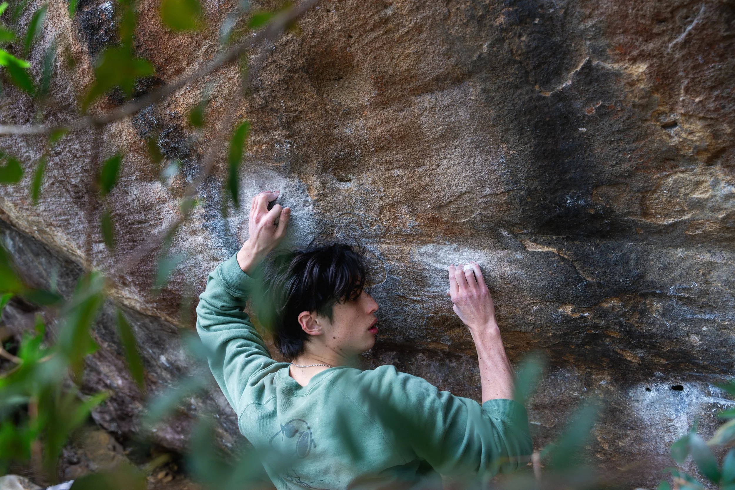 Person with short dark hair climbing a rock wall outdoors, surrounded by green foliage.