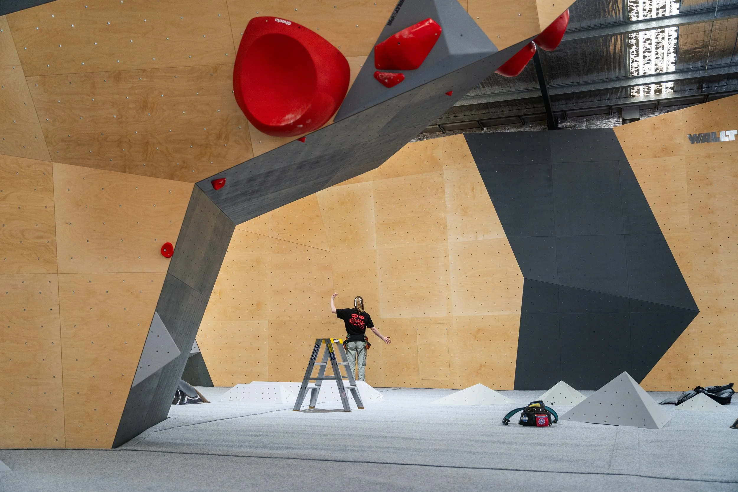 Indoor climbing gym with a large wooden climbing wall, a climber standing on the floor near the wall, and hold gear nearby.
