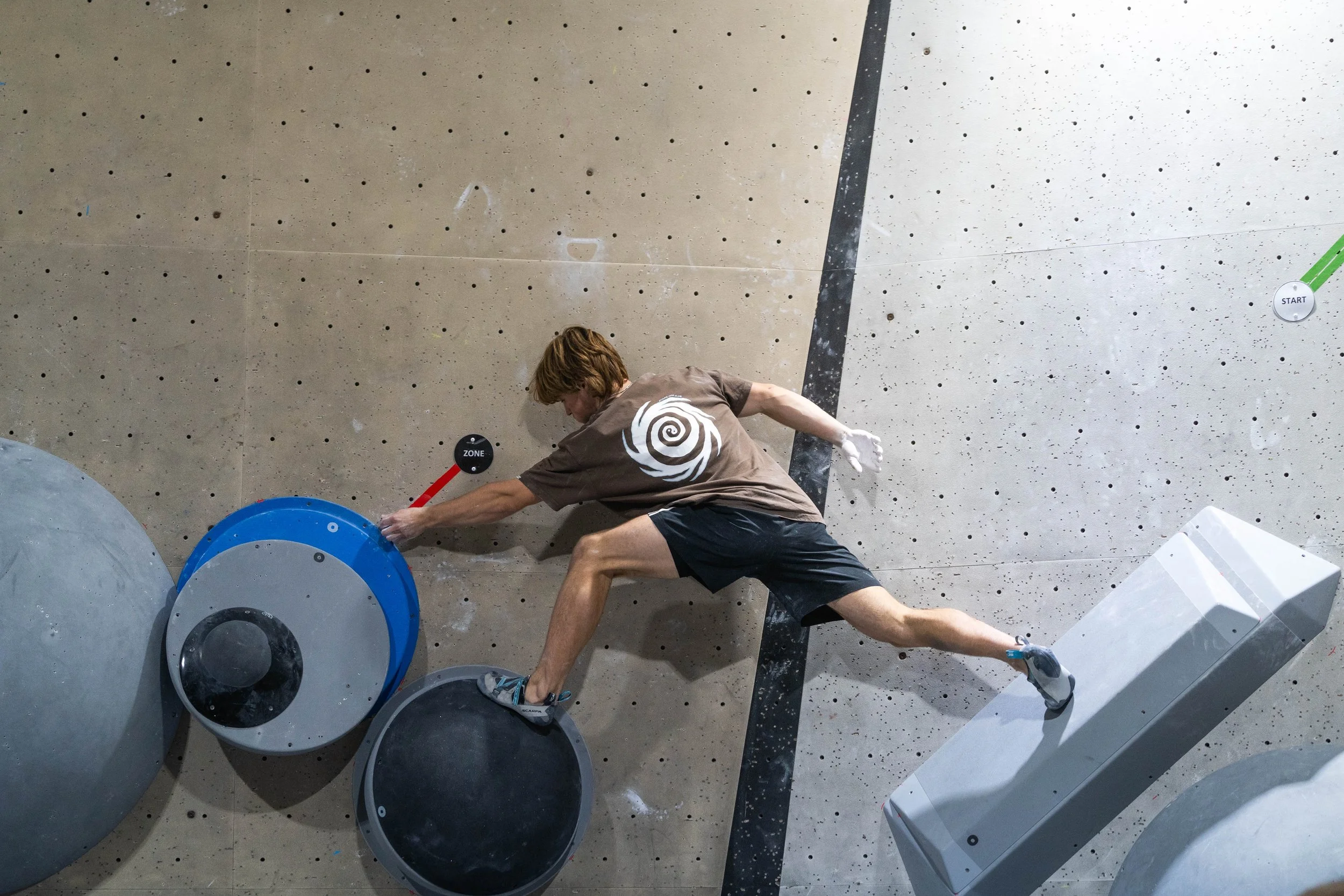 A person mid-climb on indoor bouldering wall, reaching for a hold with their right hand, wearing a brown shirt and black shorts.
