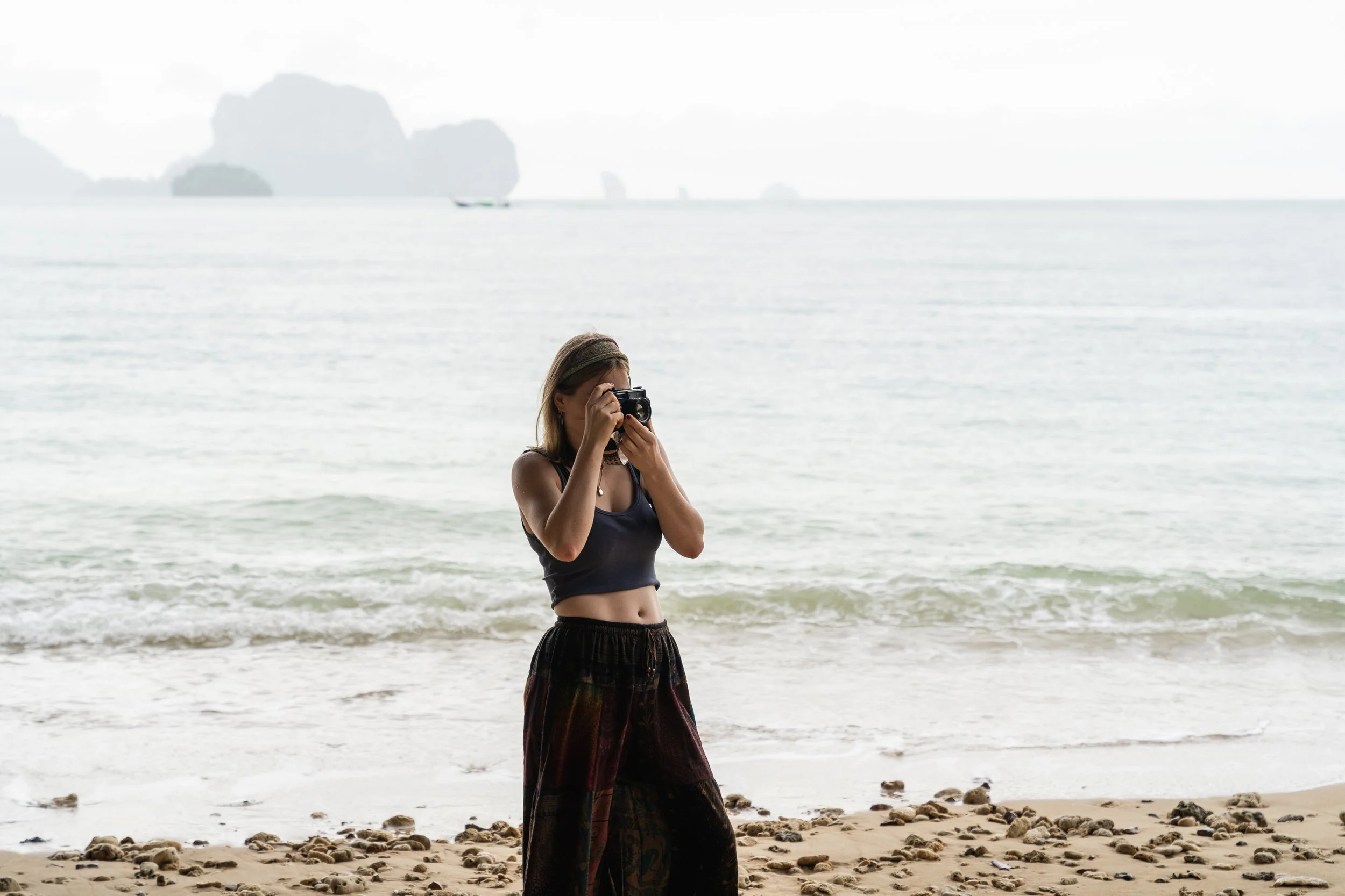 Person standing on a sandy beach with rocks, taking a photograph with a camera, ocean and distant islands in the background.